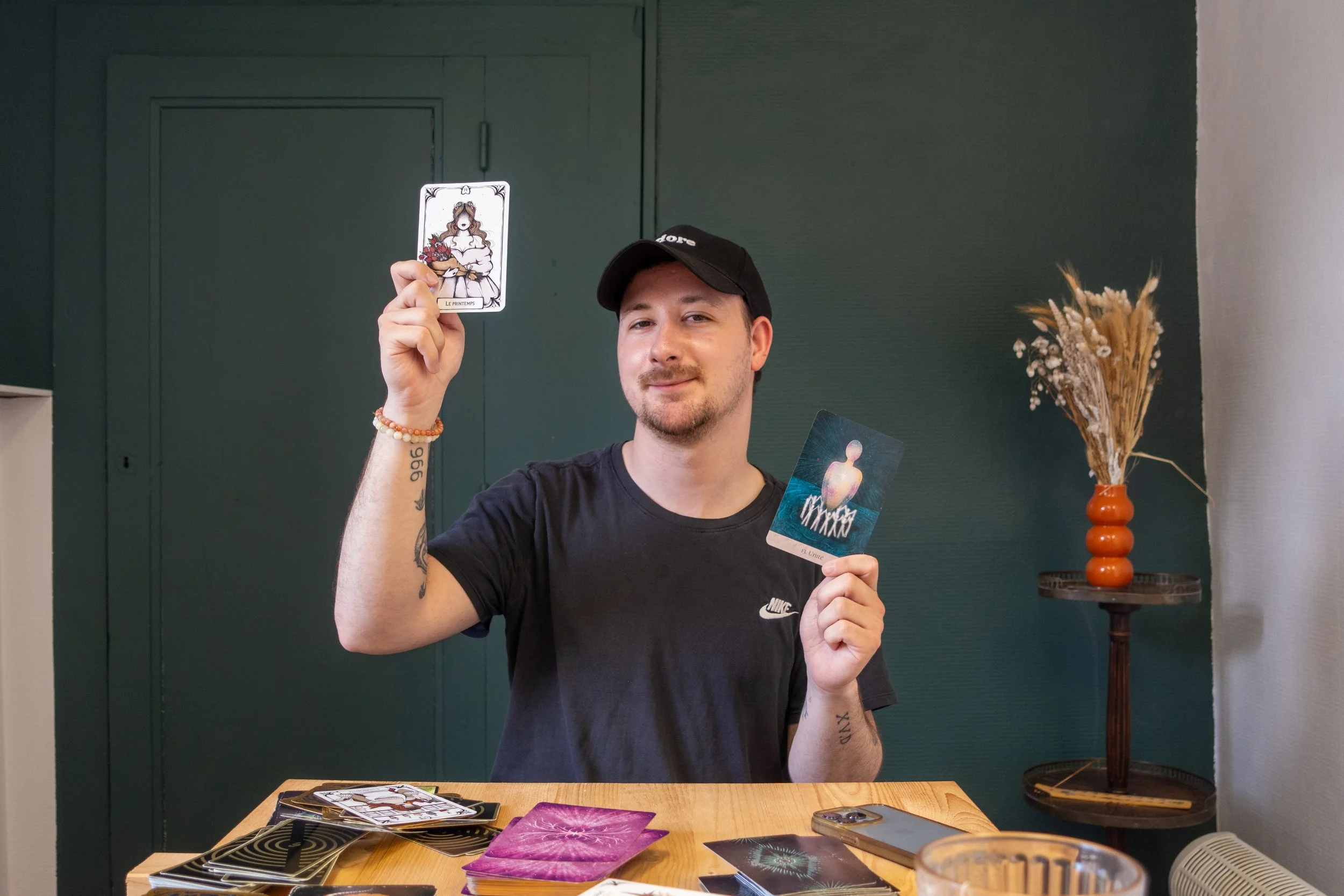Un homme assis à une table, tenant deux cartes de tarot, avec d'autres cartes posées sur la table et une plante sèche en vase à l'arrière.