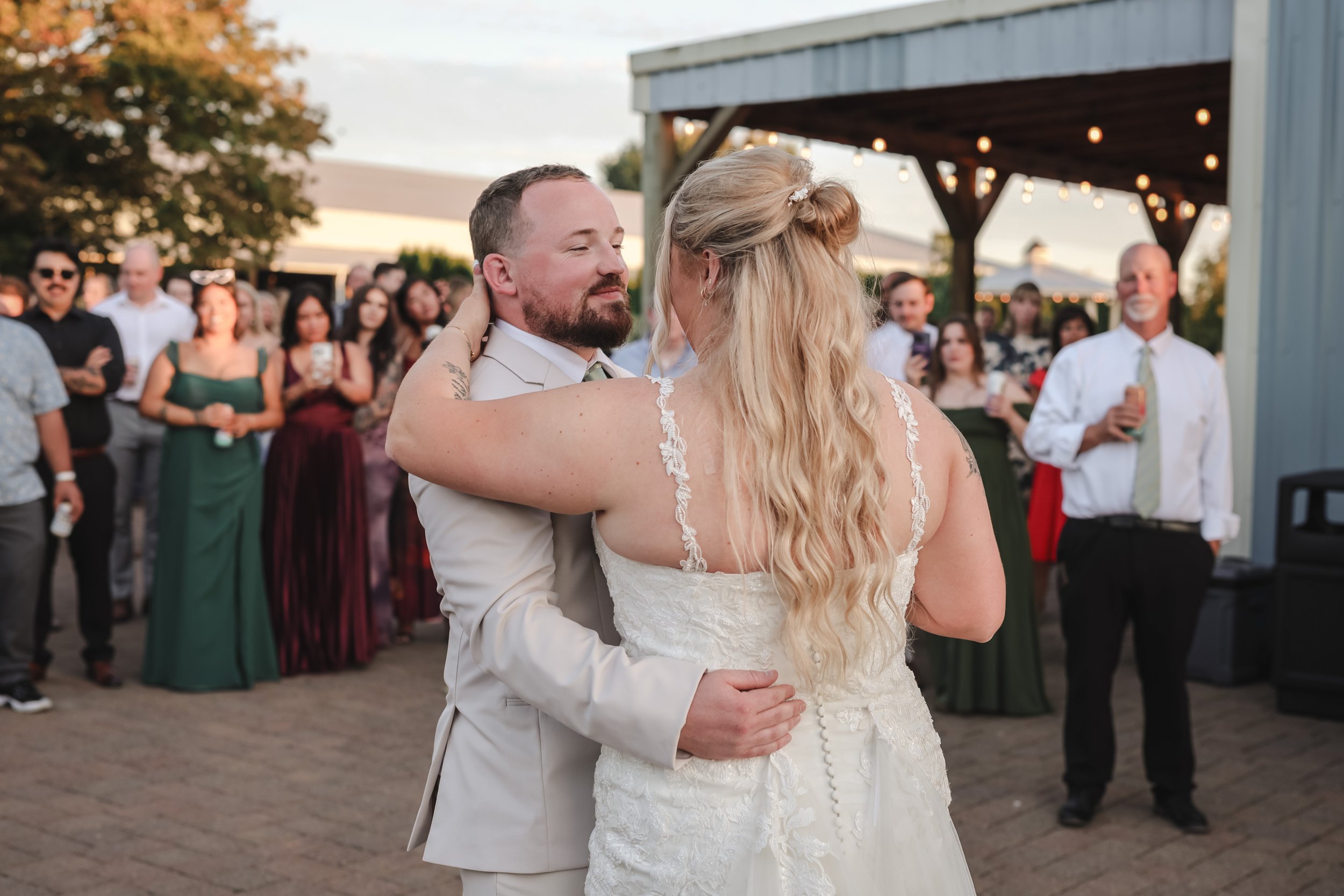 A bride and groom share a dance at their outdoor wedding reception, surrounded by guests, with string lights and trees in the background.