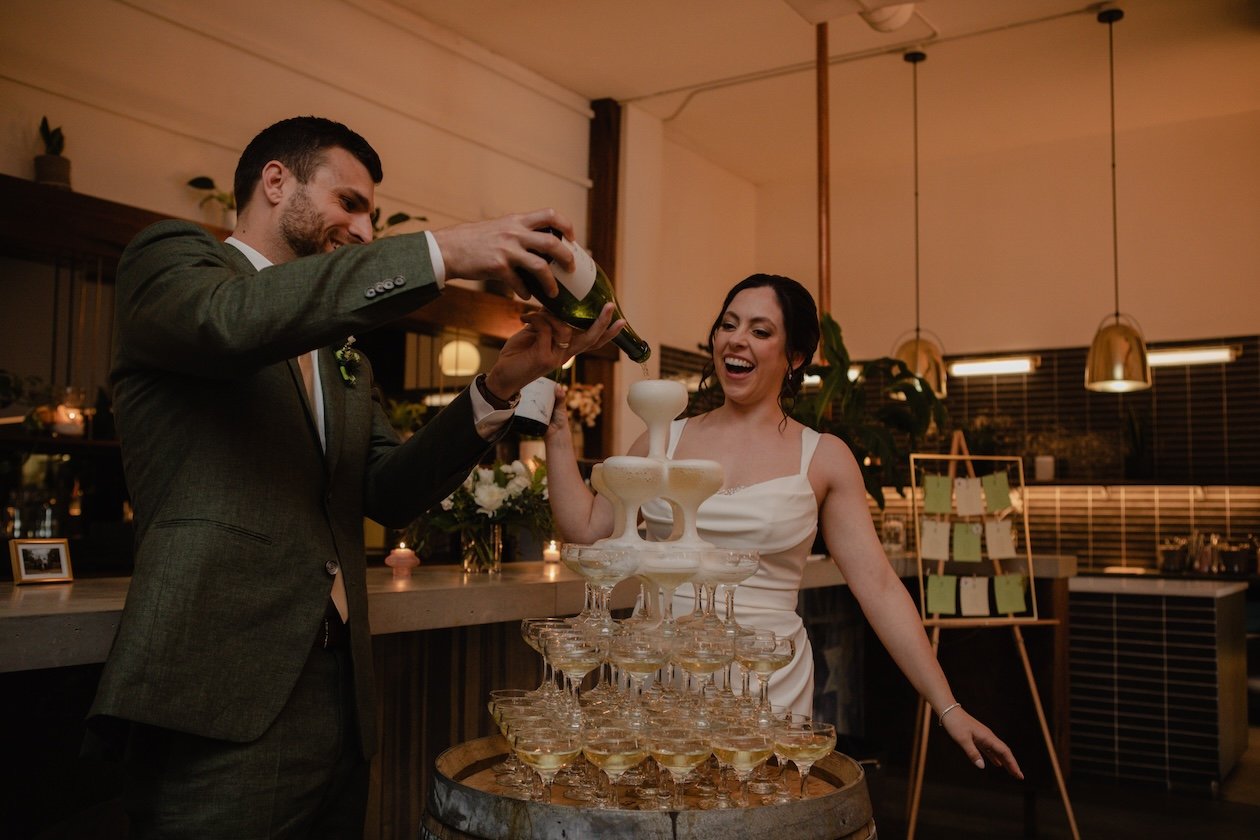 Couple pouring champagne into a glass tower at a wedding reception.