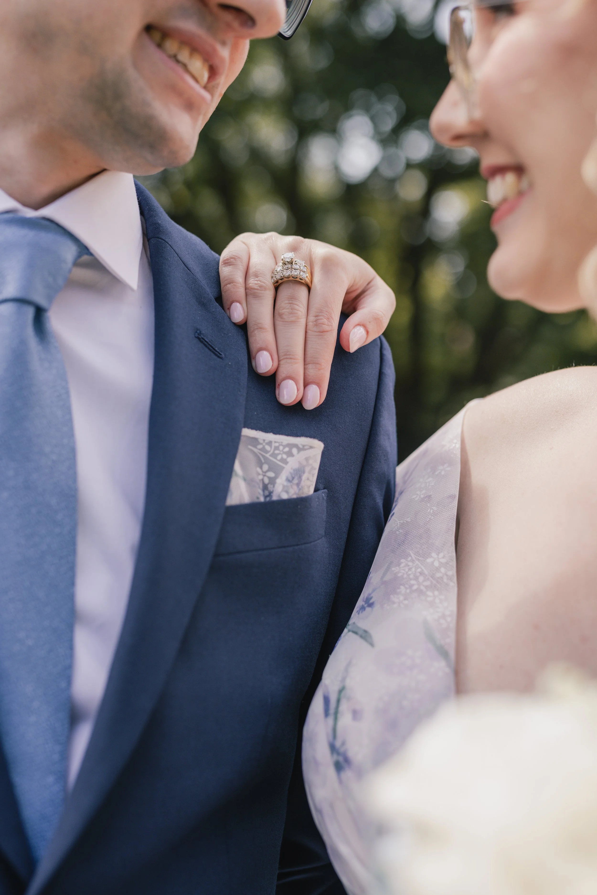 Close-up of a man in a blue suit and white shirt, smiling at a woman who is wearing a light-colored, floral-patterned dress. The woman has an engagement or wedding ring on her left hand, resting on the man's shoulder.