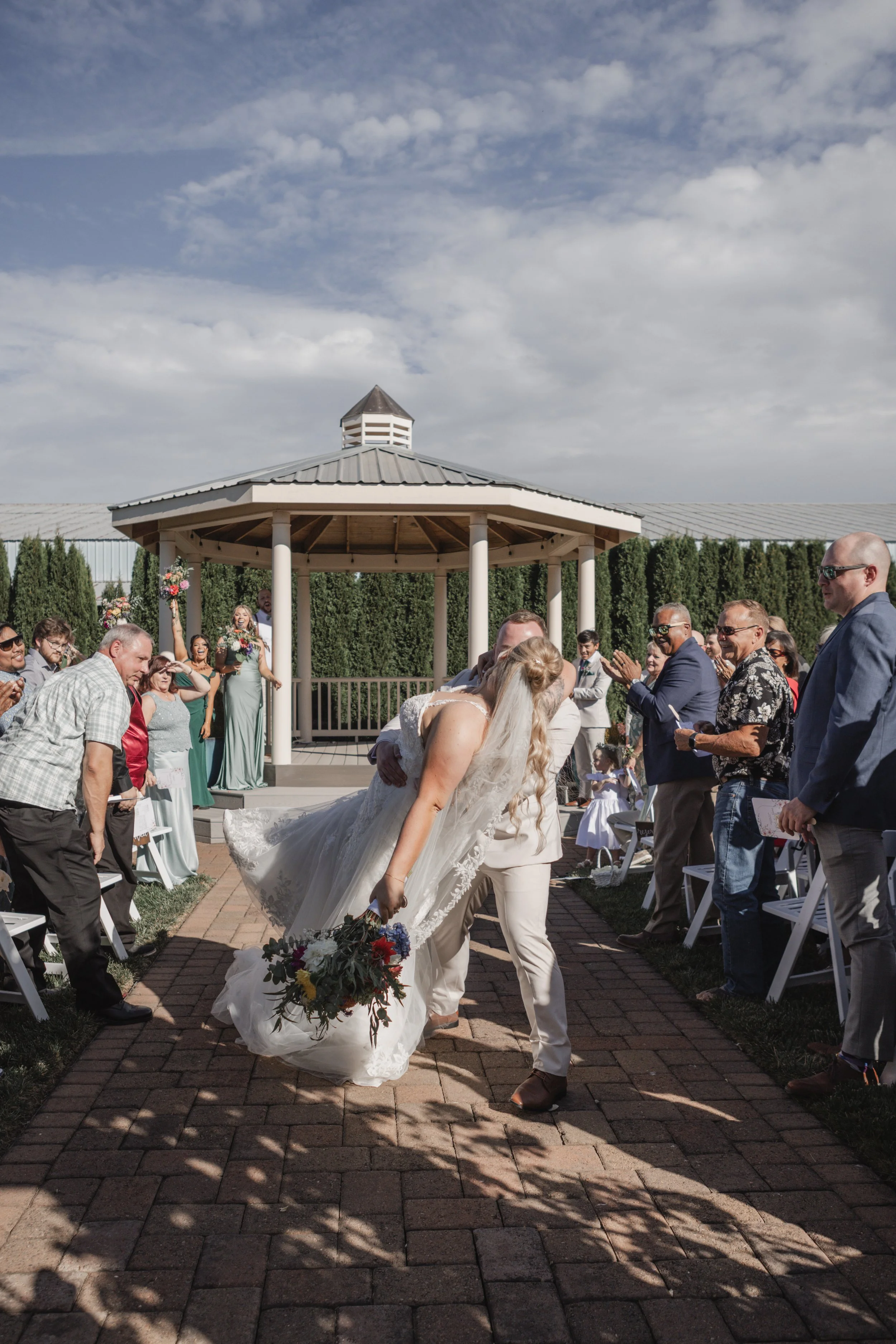 Wedding ceremony outdoors with a bride in a white dress bending down to pick up her bouquet, surrounded by guests clapping and smiling, under a gazebo on a sunny day.