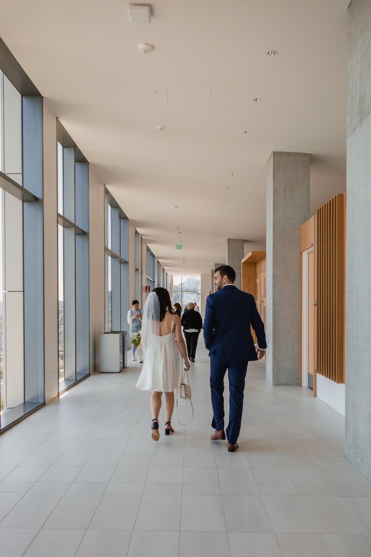 Couple walking in a modern hallway, the woman in a white dress with a veil and the man in a blue suit, large windows to the left.