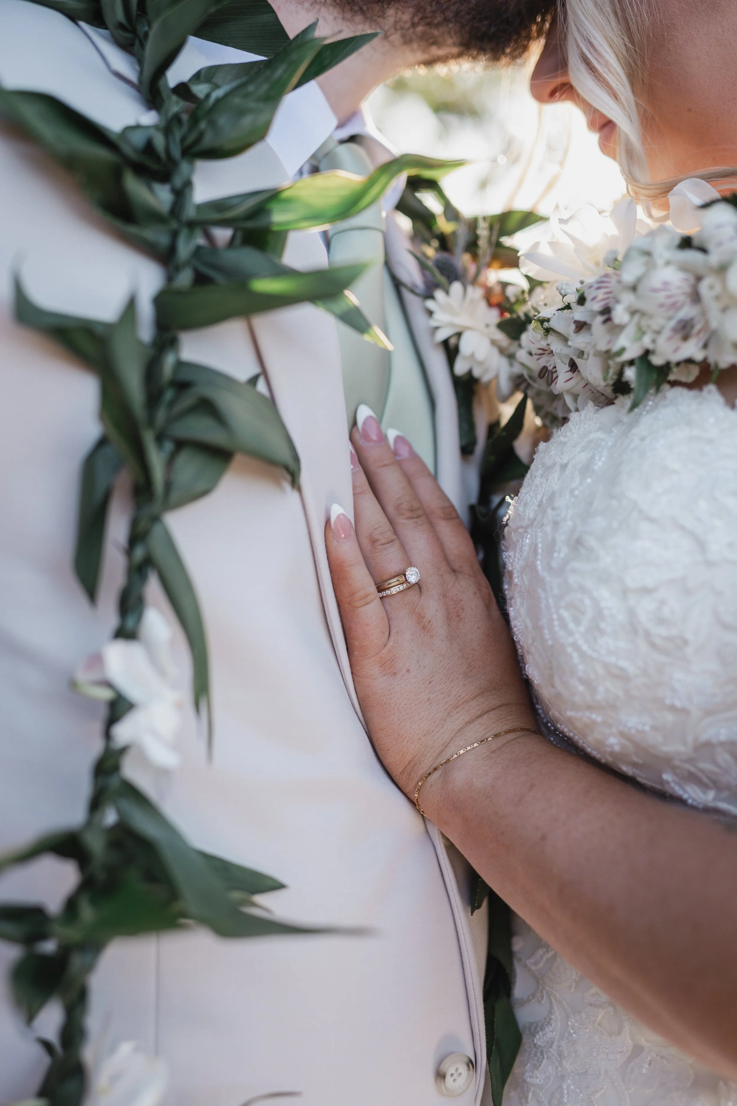 A bride and groom close-up portrait with the bride touching the groom's jacket, both embracing and wearing floral accessories, during a wedding ceremony.