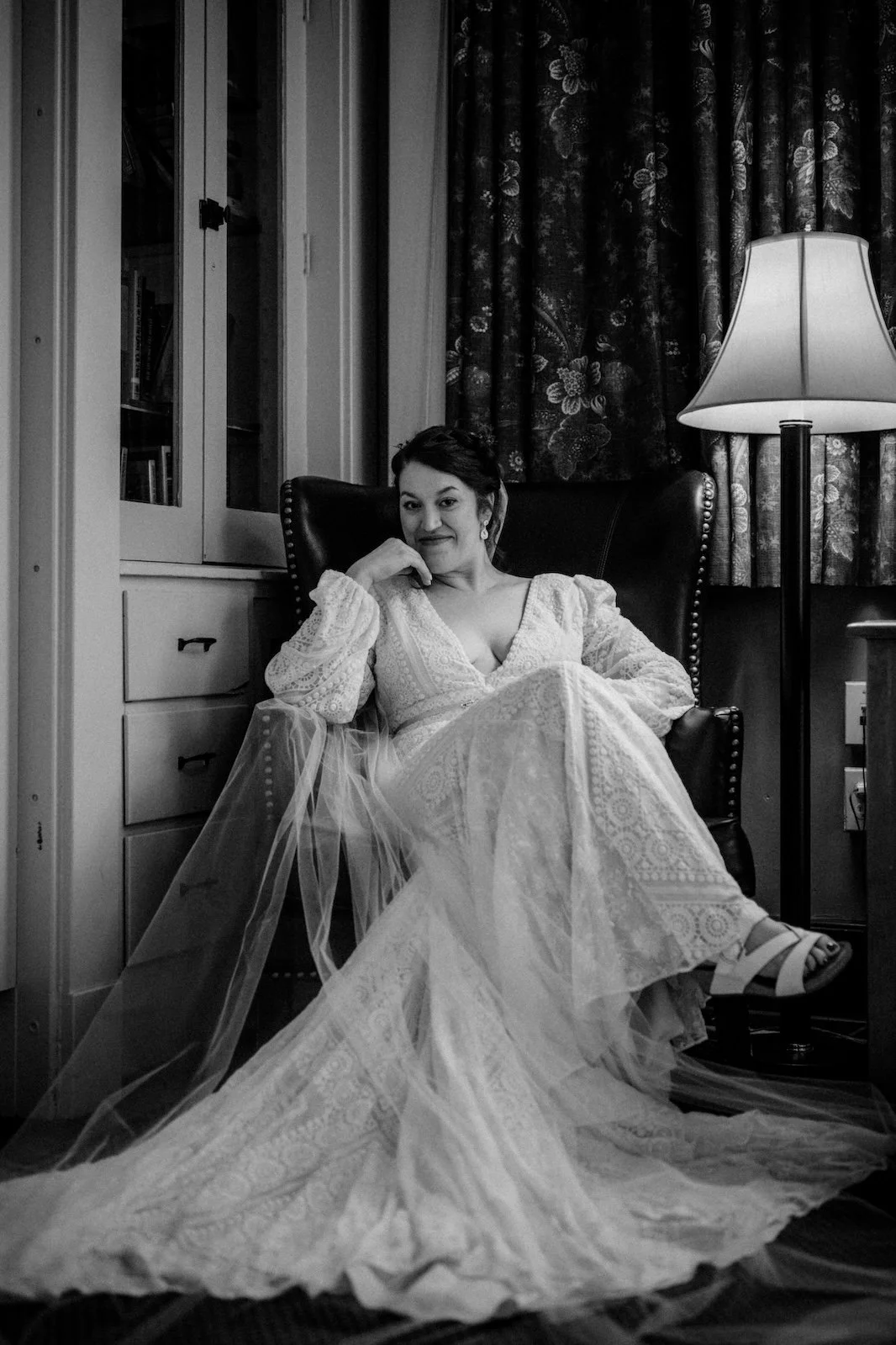 A woman in a lace wedding dress sitting in a chair next to a lamp, with floral curtains in the background.