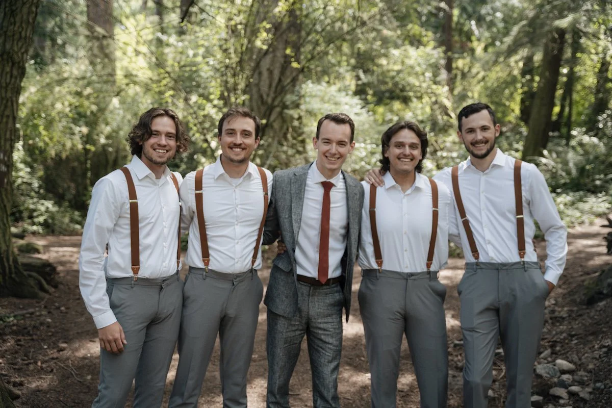 Five men posing in a forest setting, four wearing white shirts with suspenders and gray pants, one in a gray suit with a red tie.