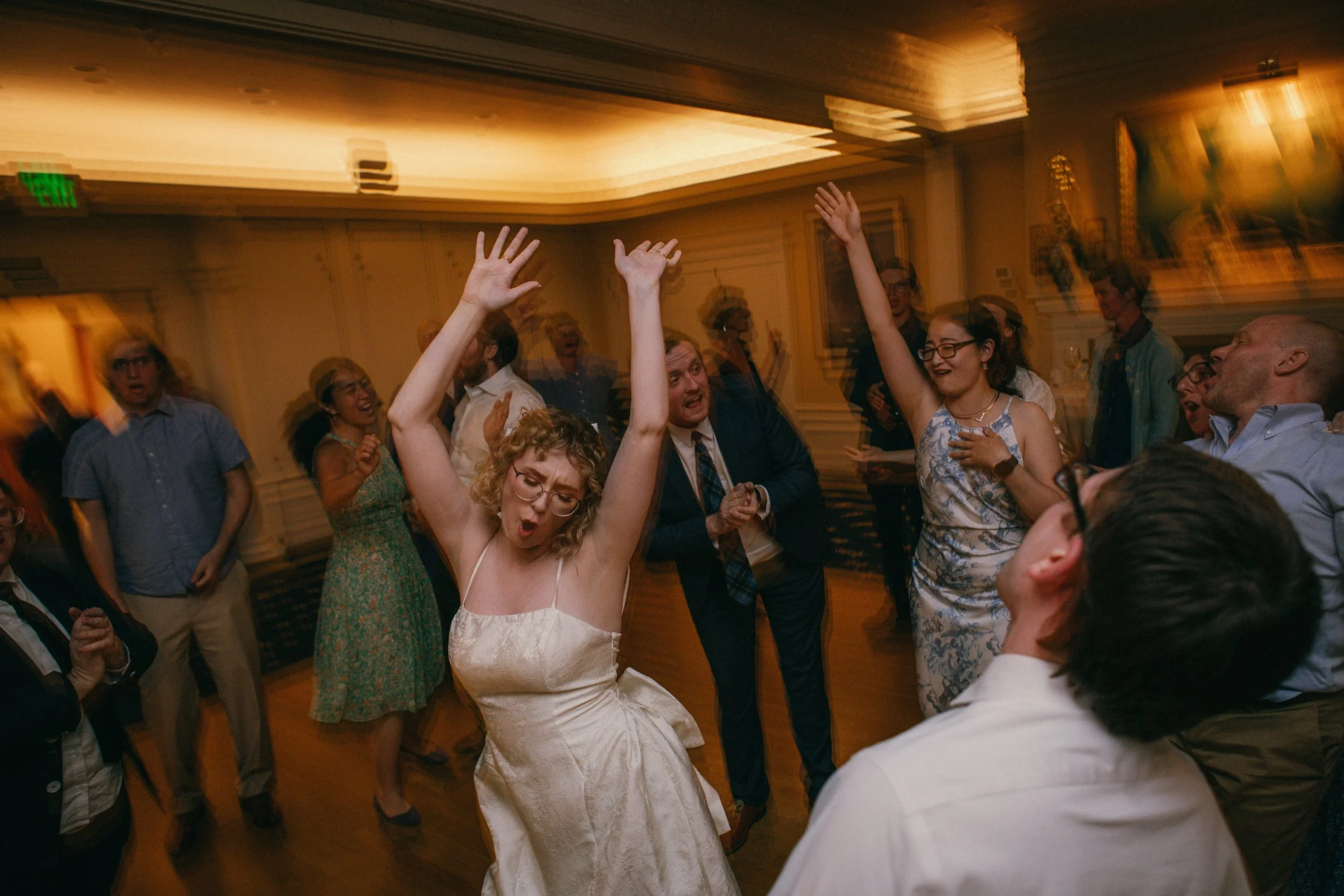 A woman in a white dress dancing and raising her hands at a lively party with other people dancing and celebrating in the background.