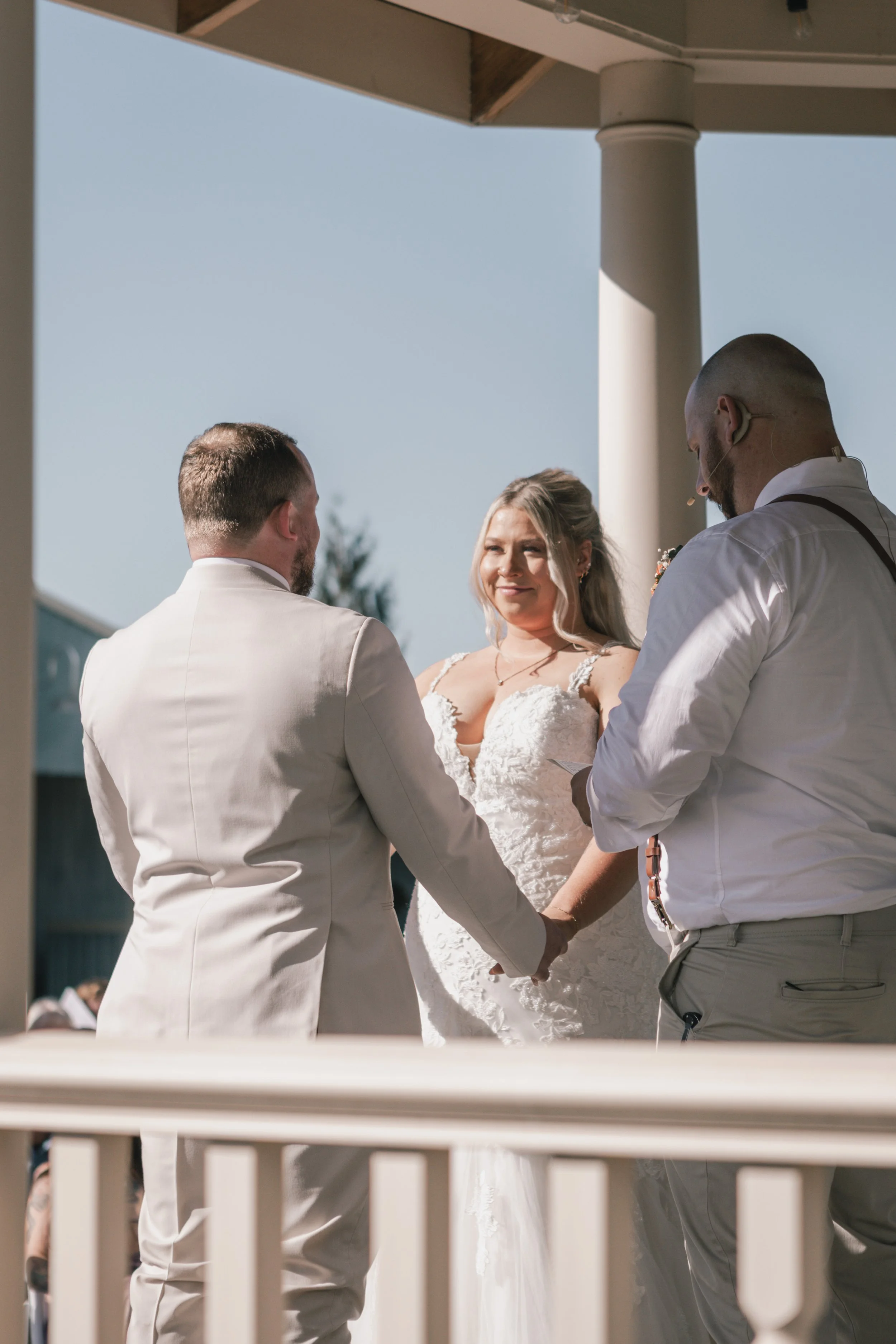 A wedding ceremony outdoors with a couple holding hands and facing each other, surrounded by friends and family. The bride is in a white lace gown, and the groom is in a beige suit. They are exchanging vows under a wooden pergola with a blue sky in t