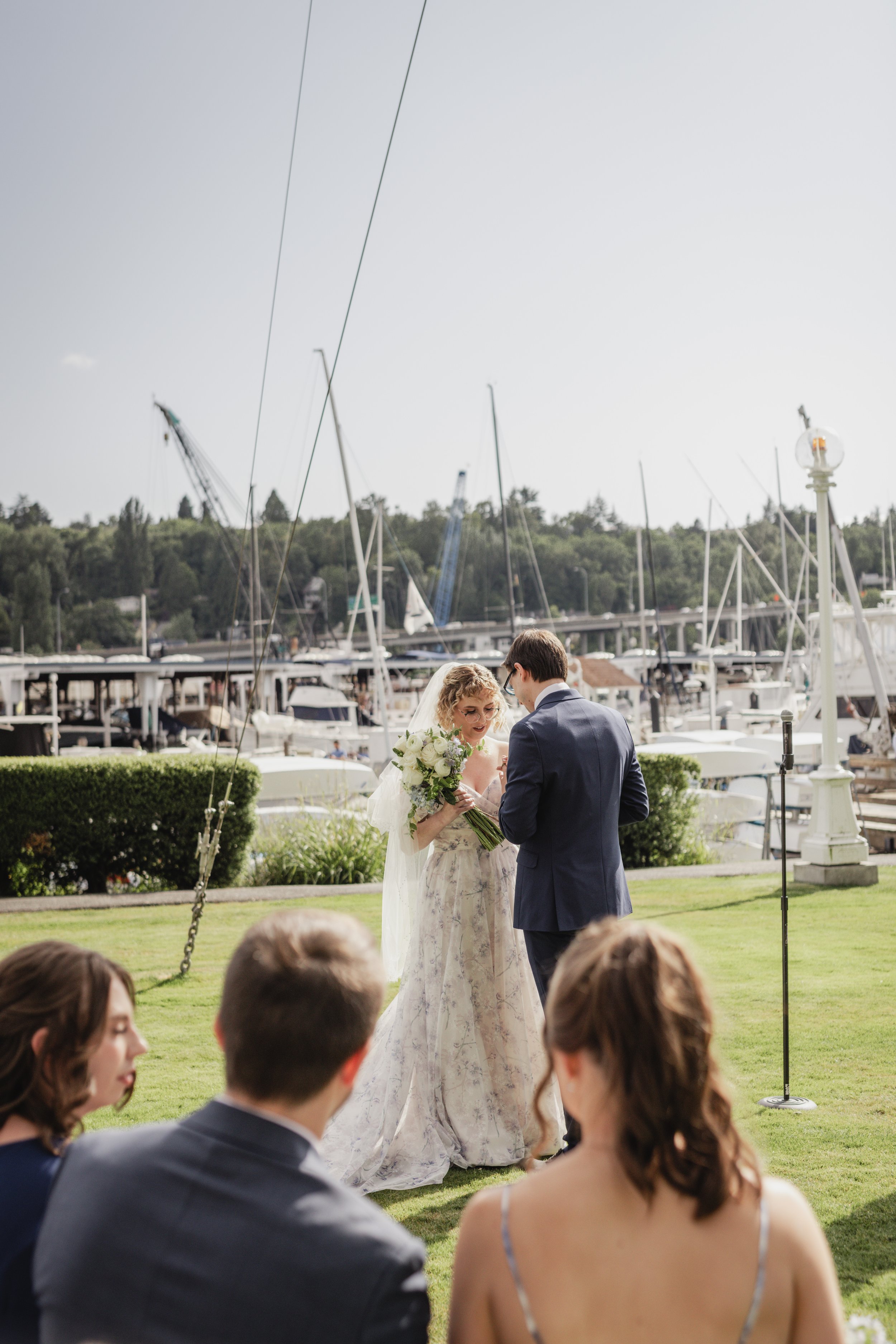 A bride and groom exchanging wedding vows outdoors at a marina, with guests facing them and sailboats docked in the background.
