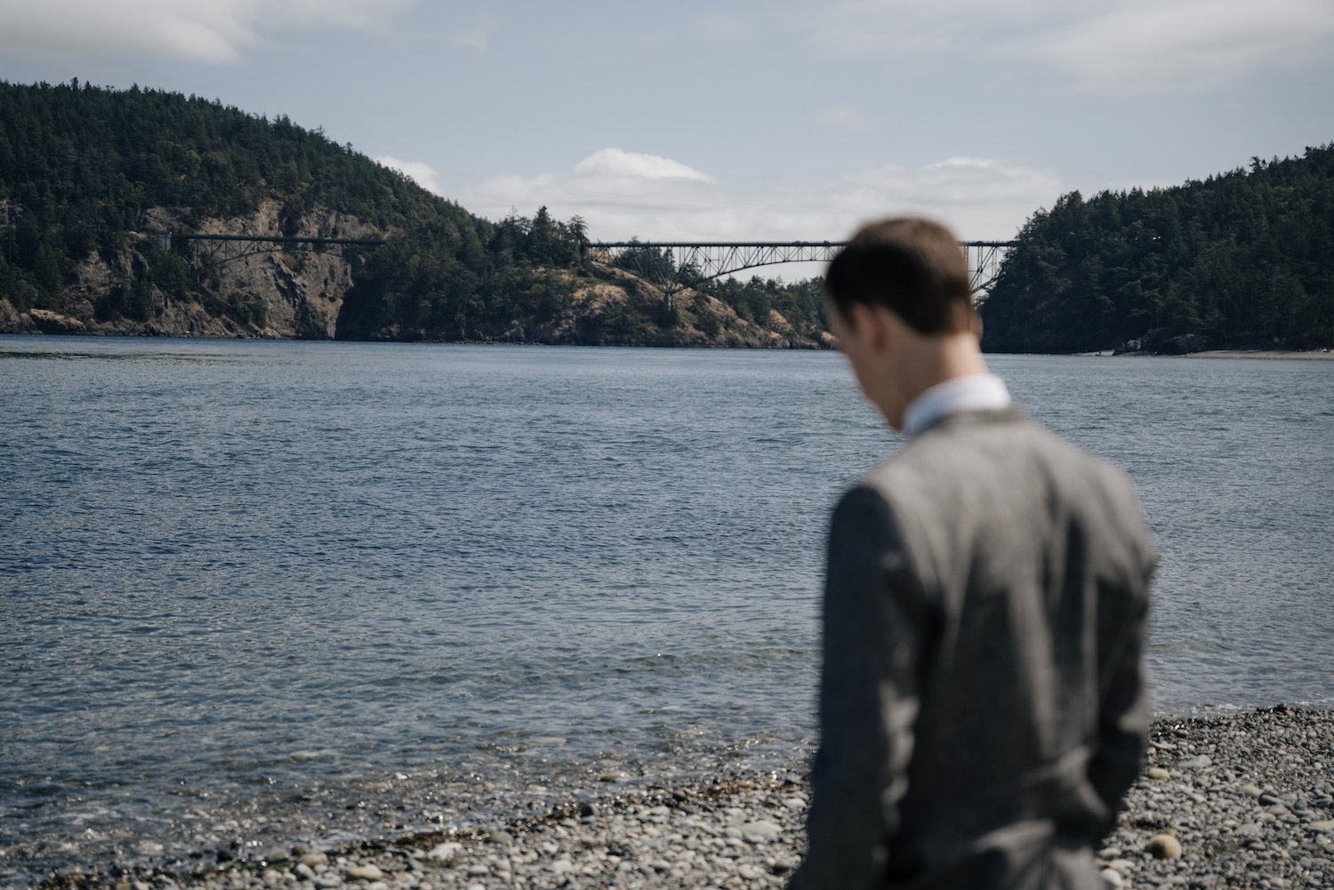 Man in suit standing on rocky beach, overlooking a bay with a distant bridge and forested hills.