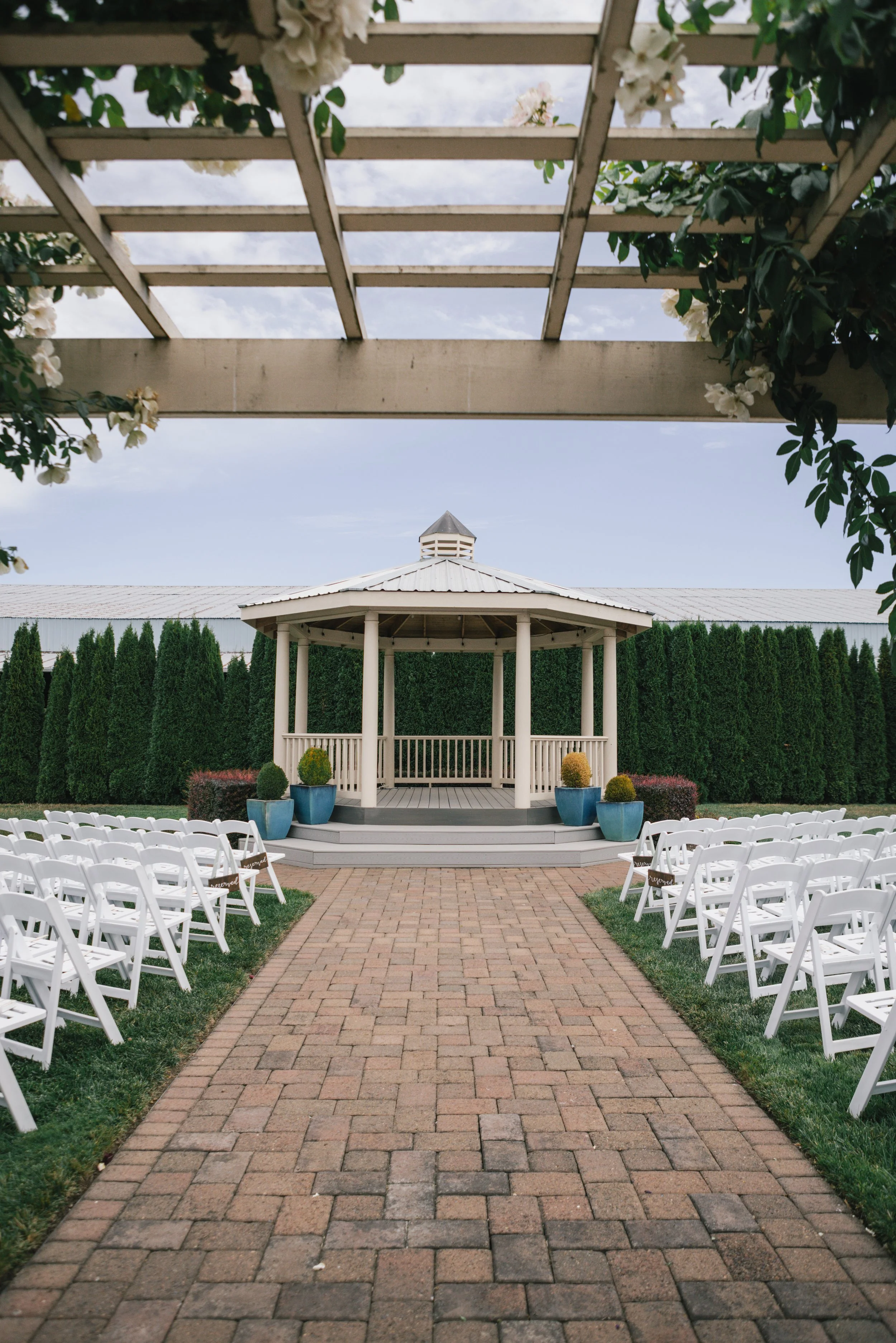 A wedding ceremony setup in an outdoor space with white folding chairs arranged on either side of a brick aisle leading to a white gazebo. The gazebo is surrounded by tall green hedges and decorated with potted plants, with a cloudy sky overhead.