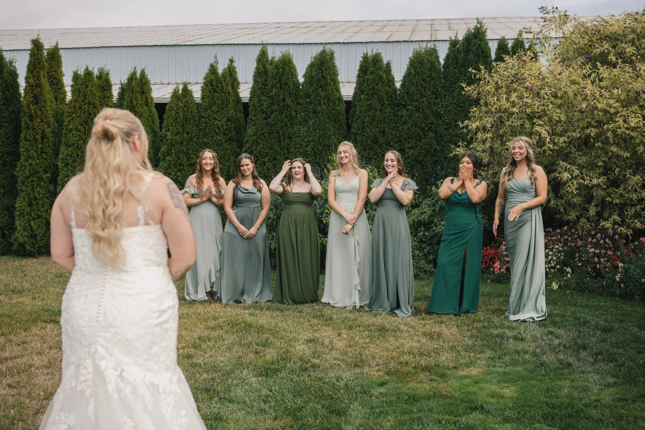 A bride in a white lace wedding dress facing six bridesmaids in light green dresses, standing outdoors on grass with trees and a white building in the background, all smiling and looking at the bride.