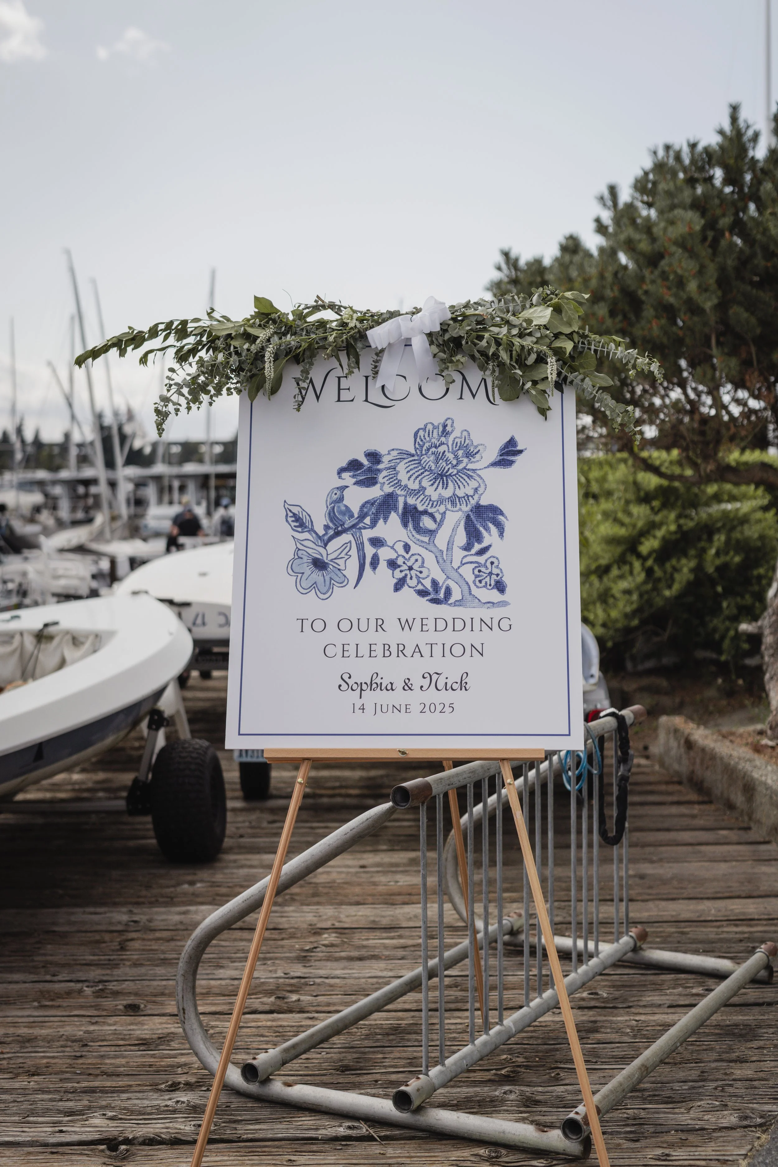 Wedding sign decorated with green foliage and a white ribbon, welcoming guests to a wedding celebration for Sophia and Nick on June 14, 2025, at a marina with boats and trees in the background.
