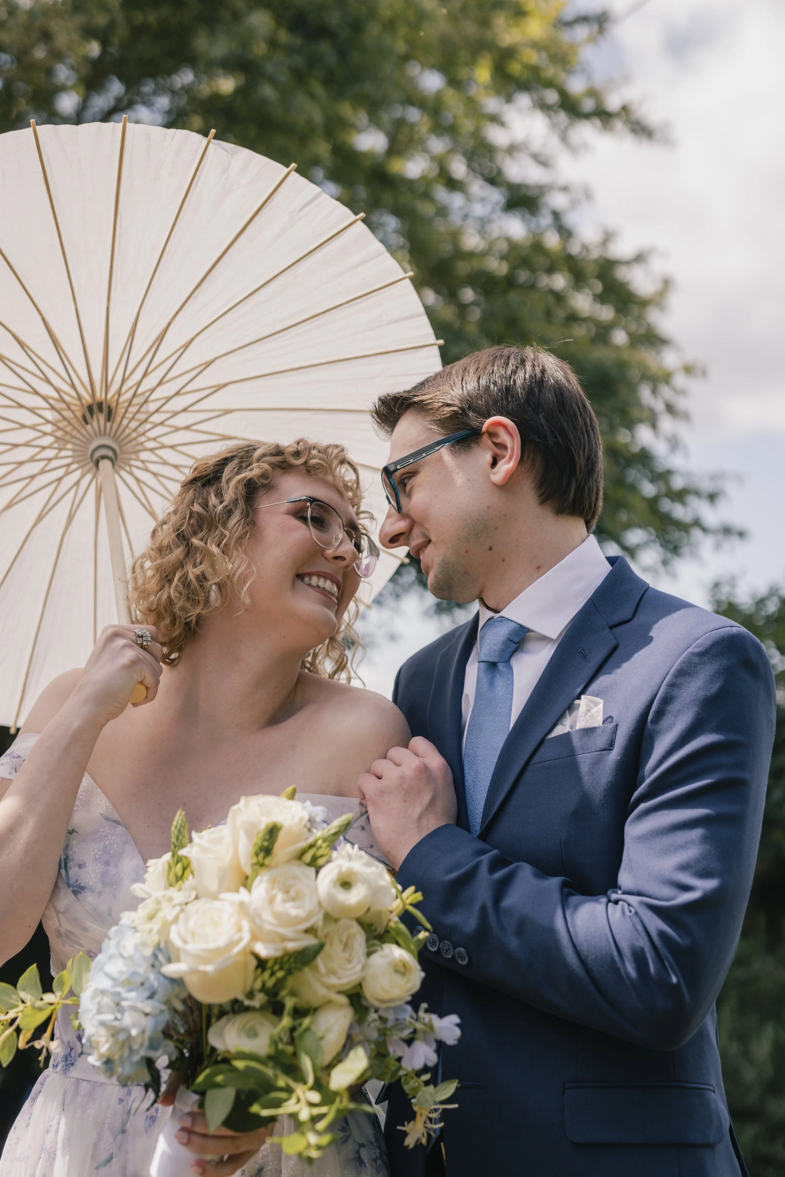 A happy bride and groom are close together, smiling and looking into each other's eyes at their wedding outdoors. The bride is holding a bouquet of white flowers and has curly blonde hair and glasses. The groom is wearing a blue suit and glasses. An 