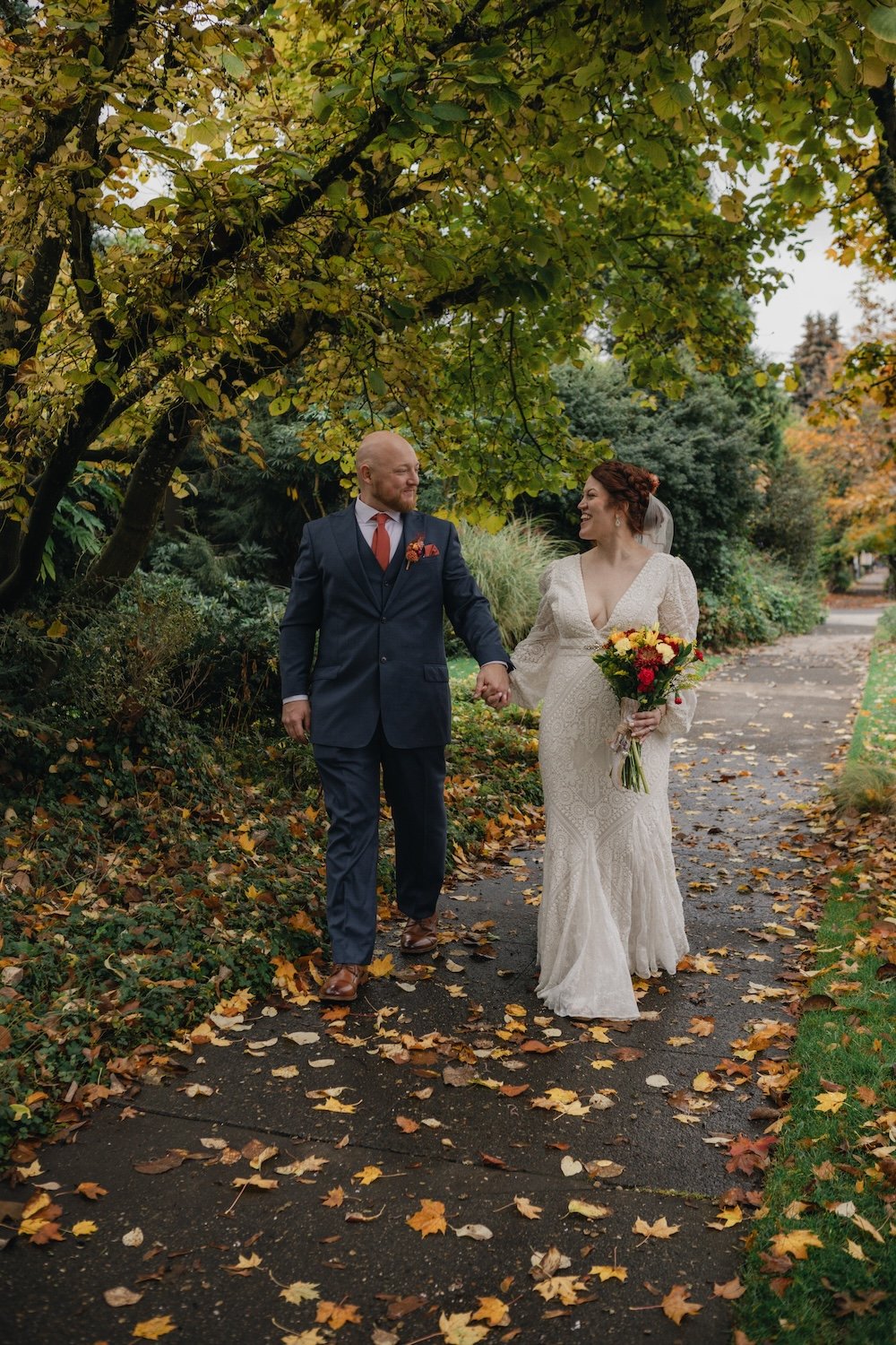 Bride and groom walking on a leaf-covered path under trees in a garden, with the bride holding a bouquet of colorful flowers.