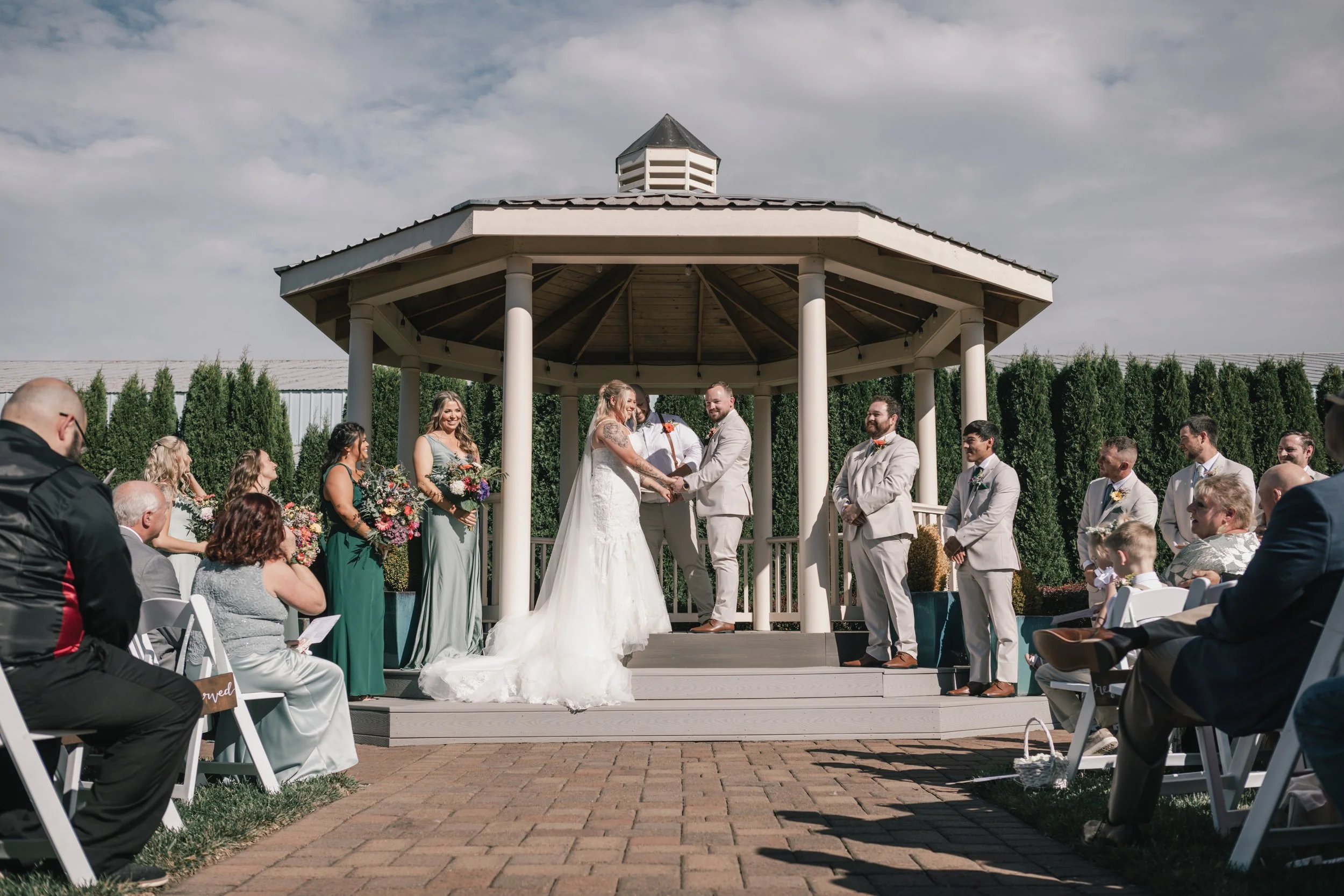 A wedding ceremony taking place outdoors under a gazebo, with the bride and groom exchanging vows, surrounded by bridesmaids, groomsmen, family, and friends seated on white chairs.