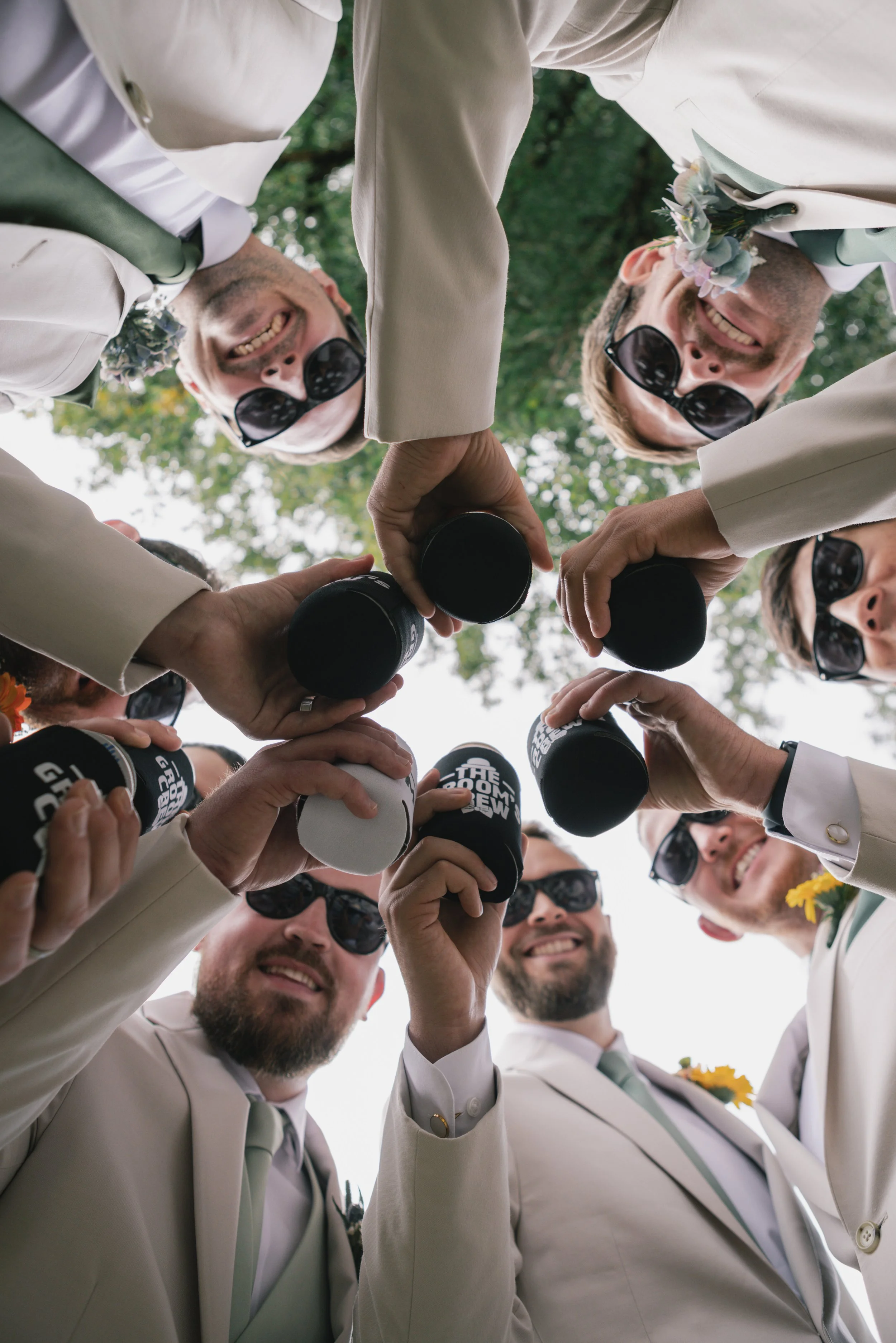 A group of men dressed in white suits and sunglasses gathered outdoors, smiling, and raising black cans together in a toast.