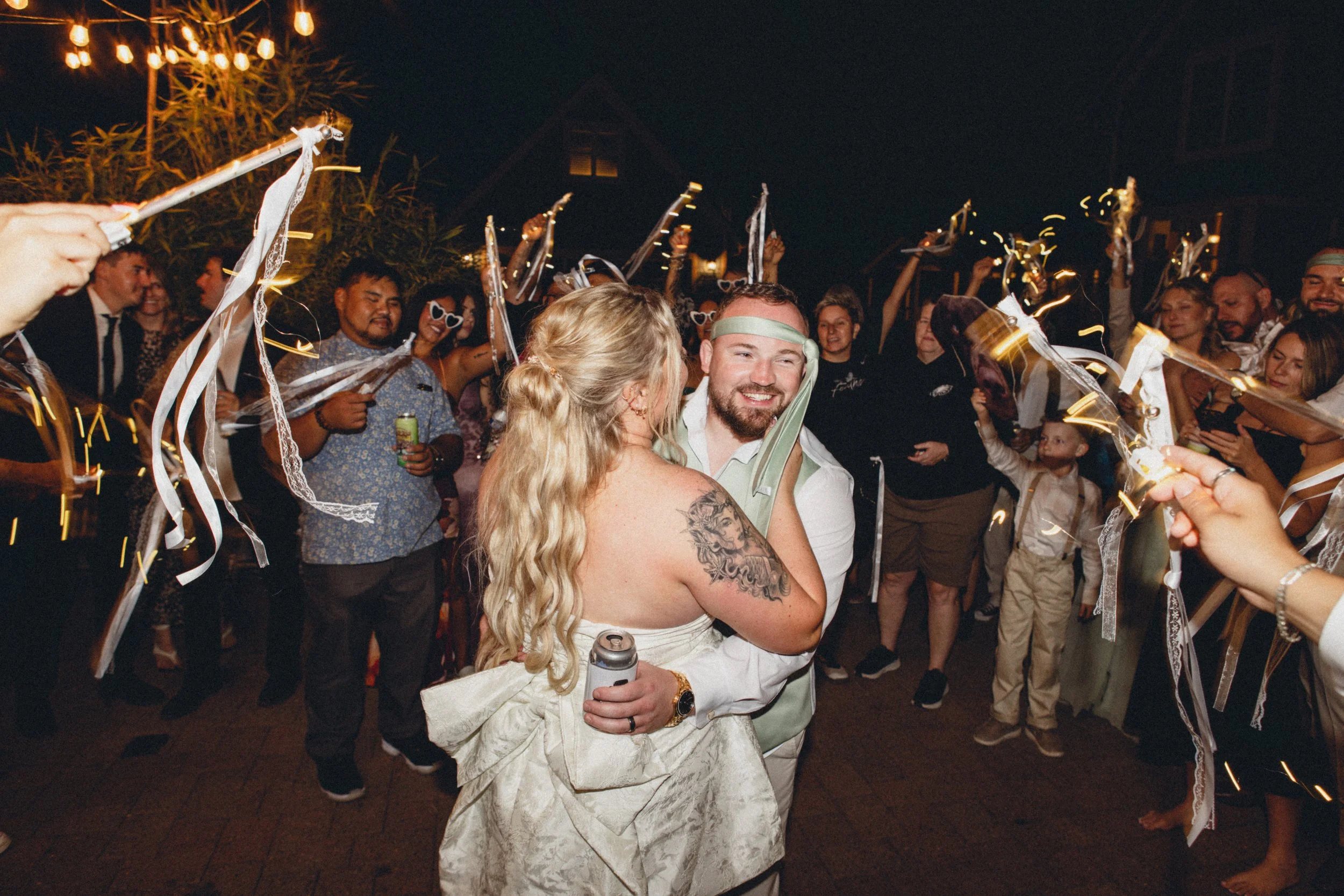 A joyous wedding celebration with the bride and groom among a lively crowd at night, holding sparklers and smiling.