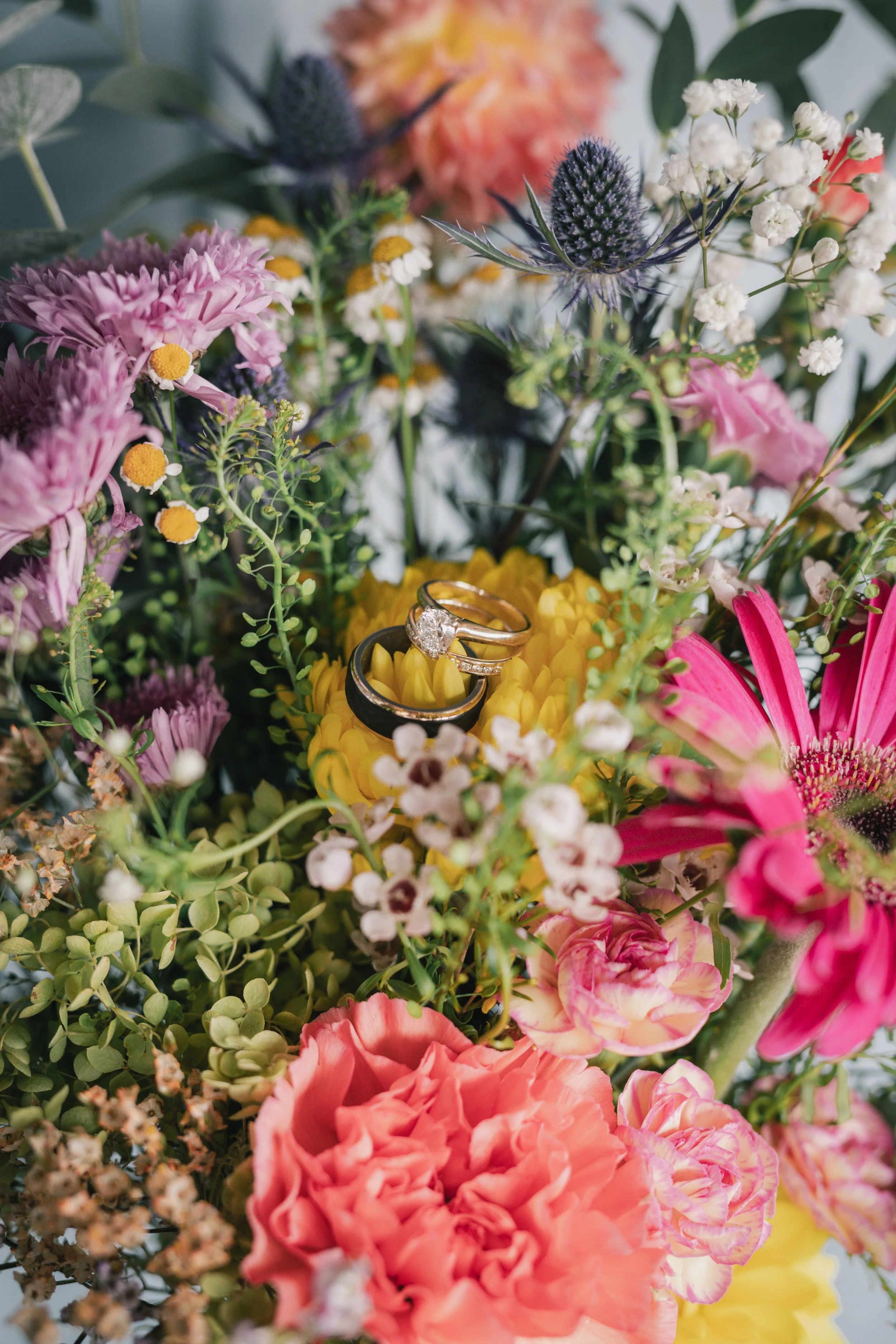 Close-up of a colorful bouquet of mixed flowers with rings placed on top, including pink carnations, purple thistles, white baby's breath, and green foliage.