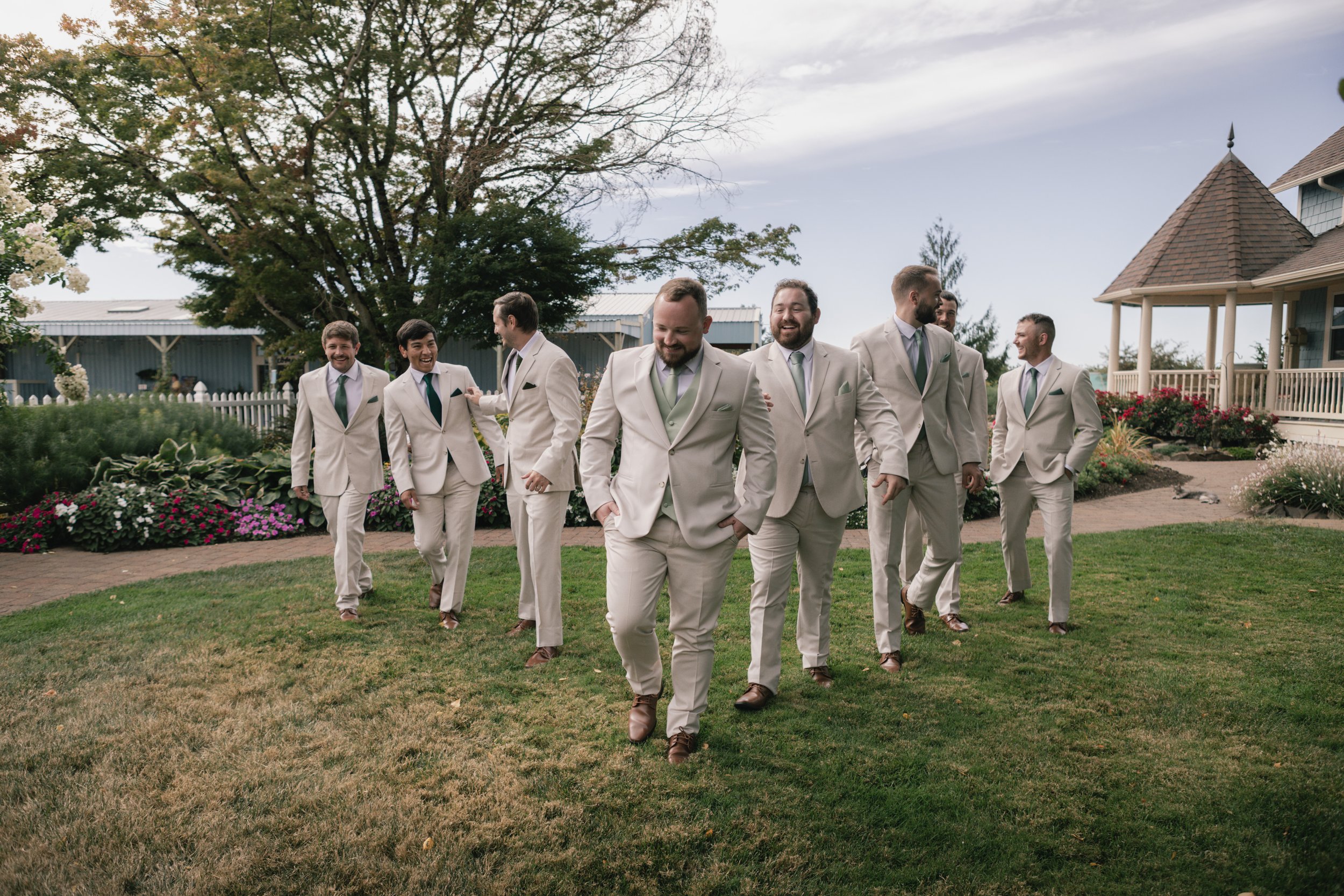 A group of men in beige suits with green ties walking on a grassy area, with a house, trees, and flowers in the background, smiling and enjoying each other's company.