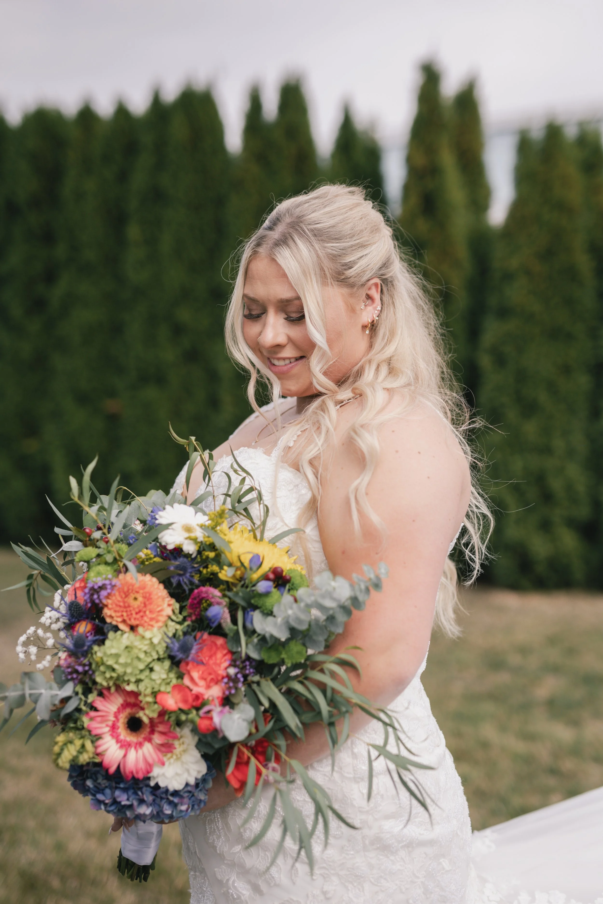 A smiling blonde woman in a white wedding dress holding a colorful bouquet of flowers outdoors, with green trees in the background.