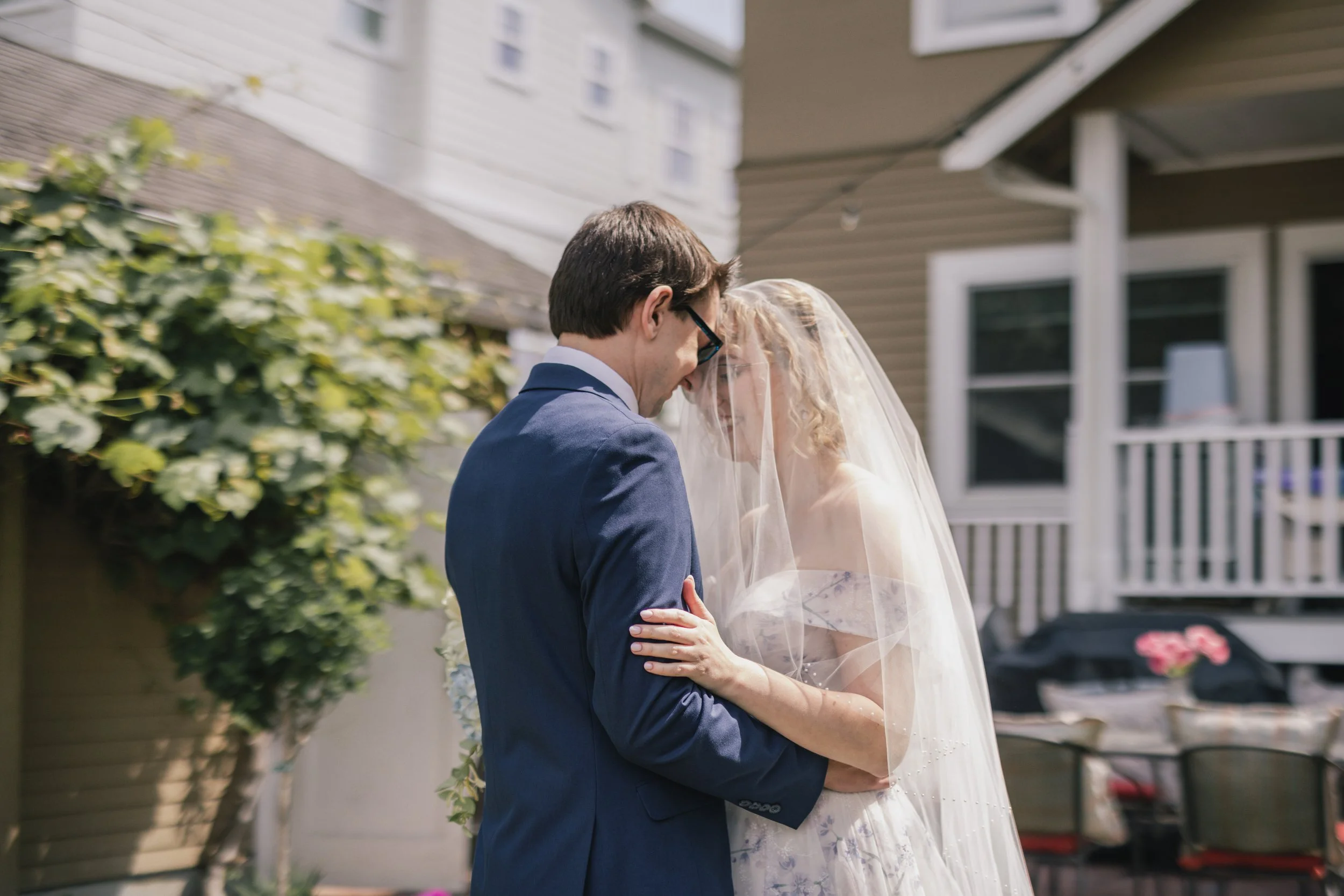 A bride and groom embracing outdoors with their foreheads touching, the groom in a blue suit and the bride in a white wedding dress with a veil, surrounded by a garden with greenery and a house in the background.