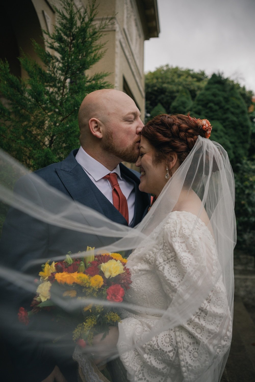 Bride and groom embracing outdoors, groom kissing bride's forehead, bride holding colorful bouquet, surrounded by greenery.