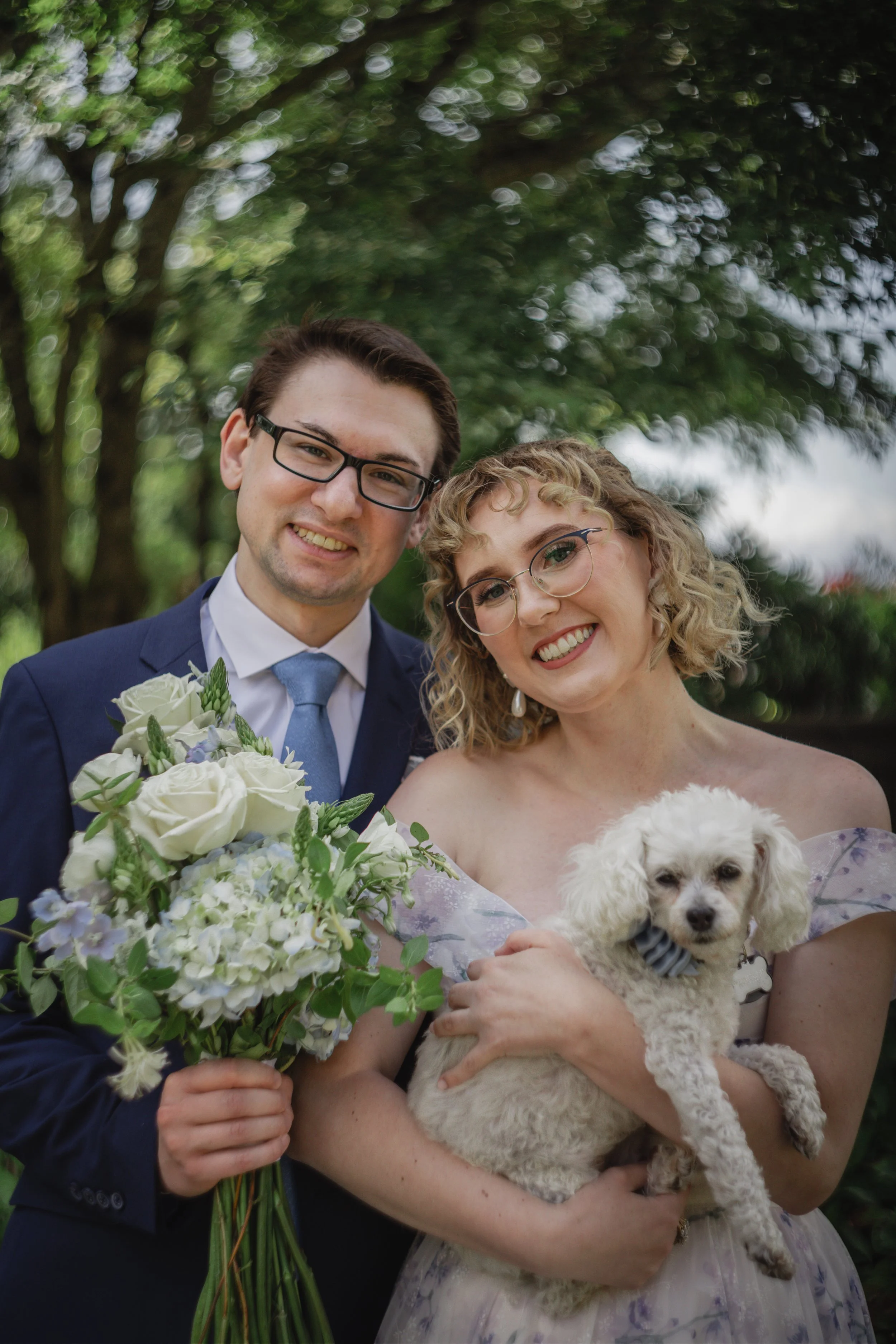 A happy couple, a man in a suit and glasses and a woman in a floral dress, standing outdoors with greenery in the background. The woman is holding a small white dog and the man is holding a bouquet of white and purple flowers.