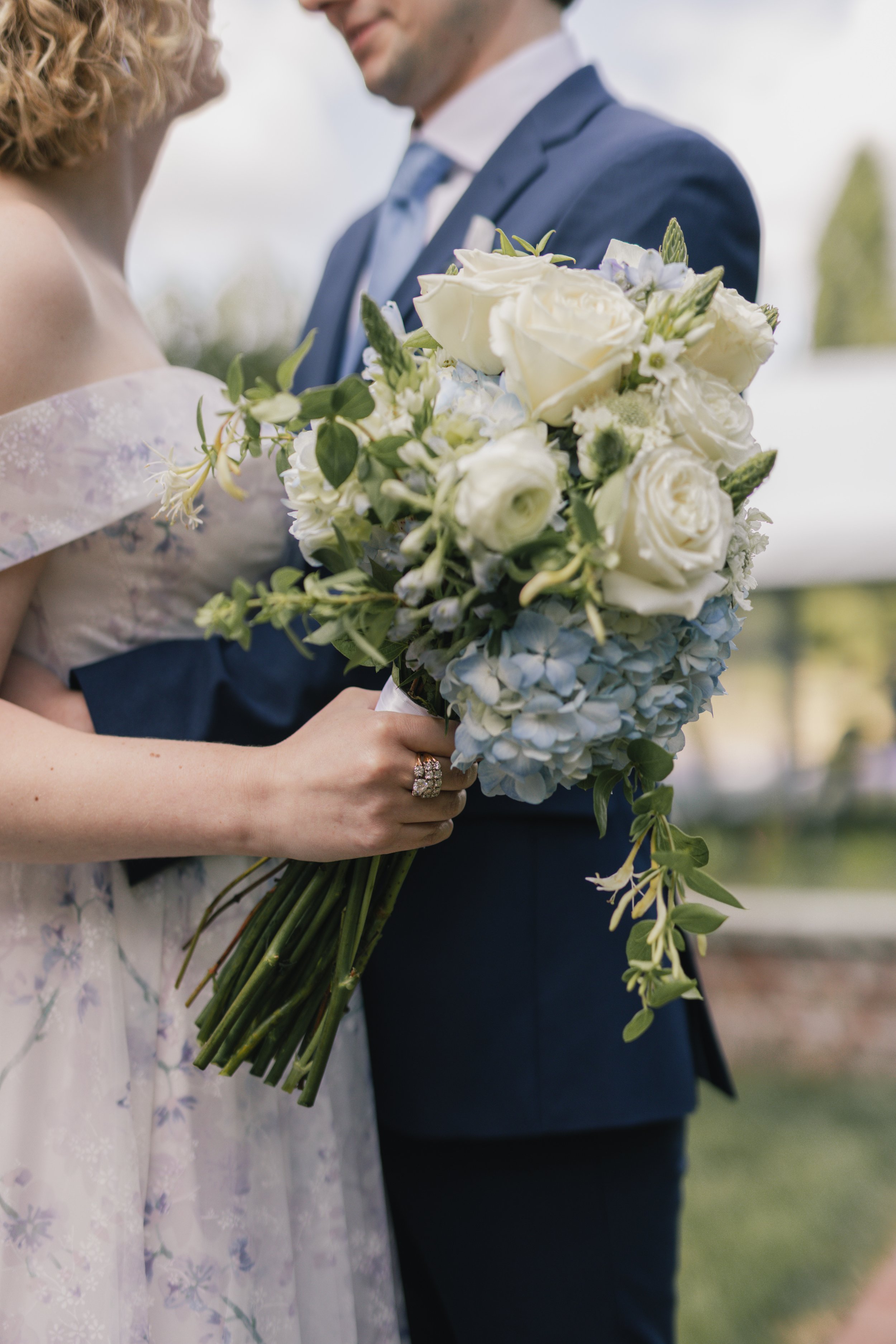 A bride holding a large bouquet of white roses and light blue hydrangeas during her wedding, standing close to a groom dressed in a navy blue suit outdoors.