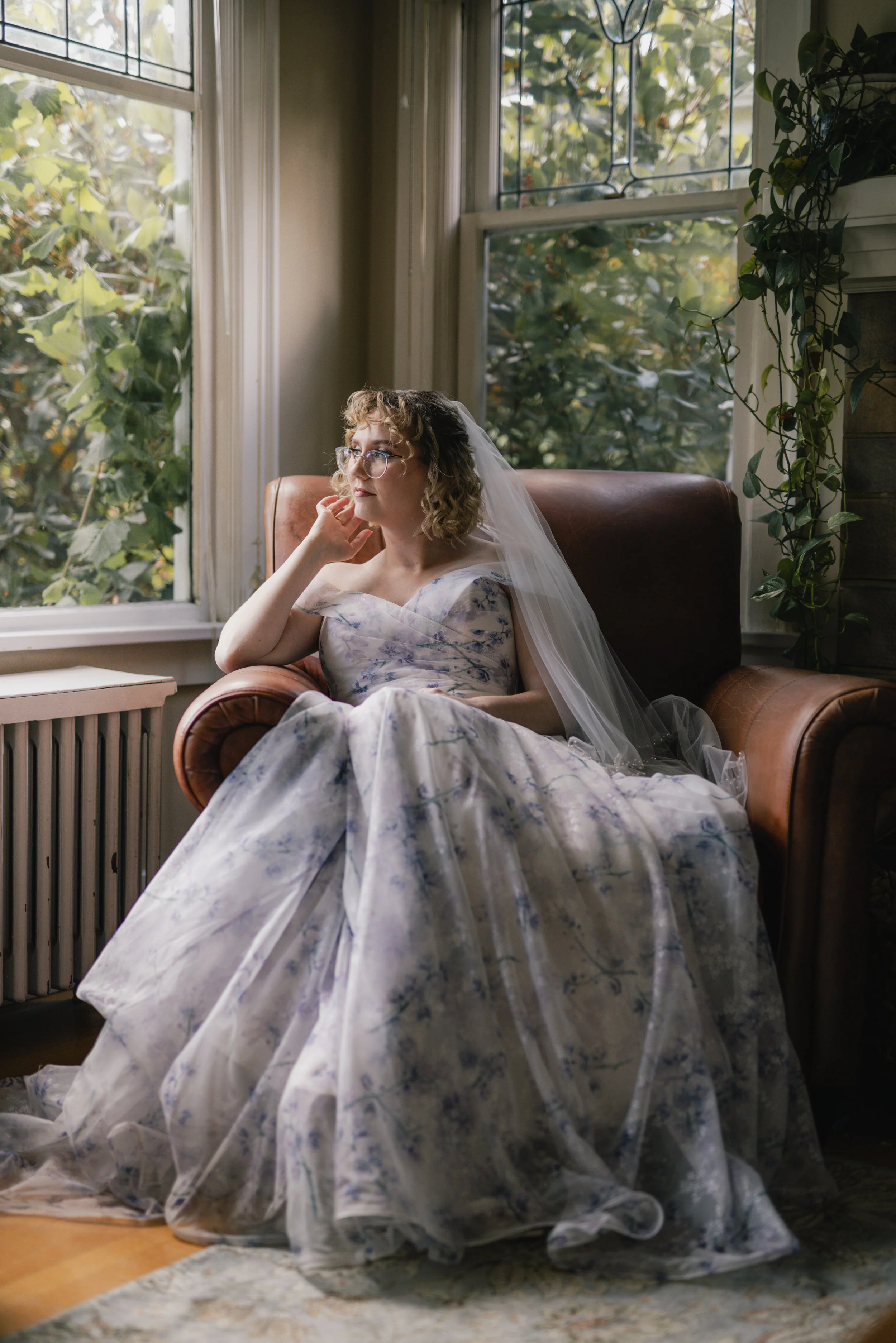 A woman with curly hair and glasses, wearing a wedding dress and veil, sitting on a leather chair near a large window with green foliage outside, inside a house.