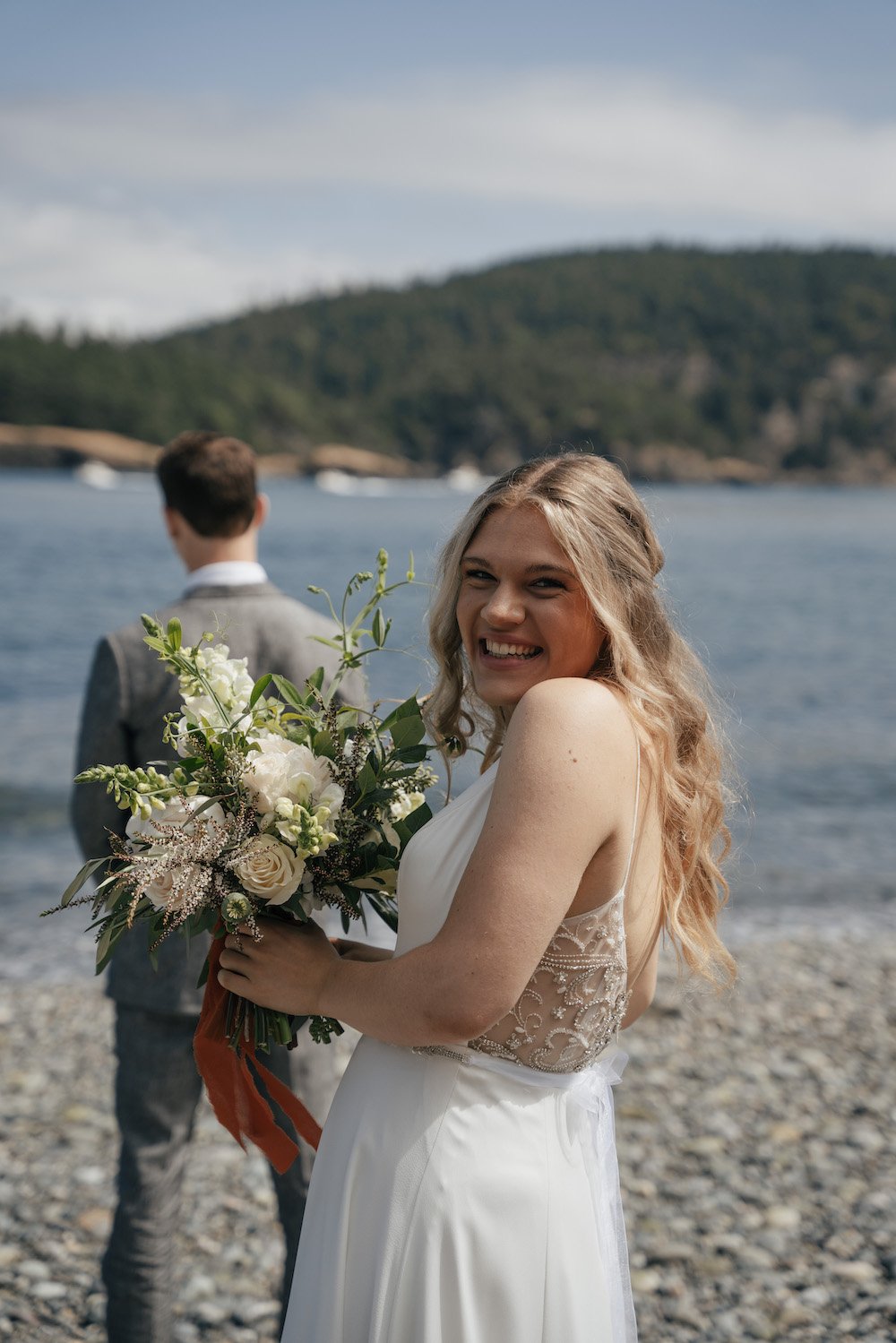 Smiling bride holding bouquet near water, groom in background.
