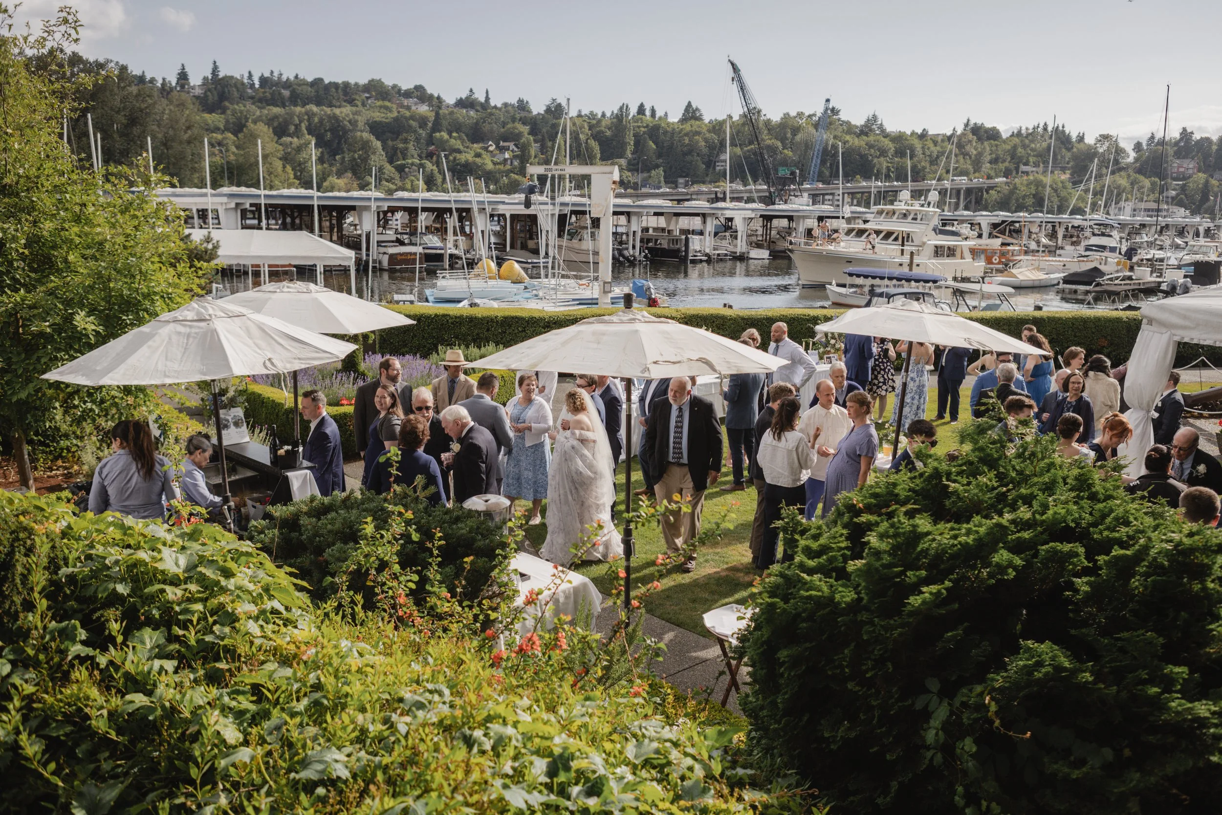 A group of people gathered outdoors at a waterfront location with boats and yachts docked, enjoying a social event under large white umbrellas and surrounded by green landscaping.