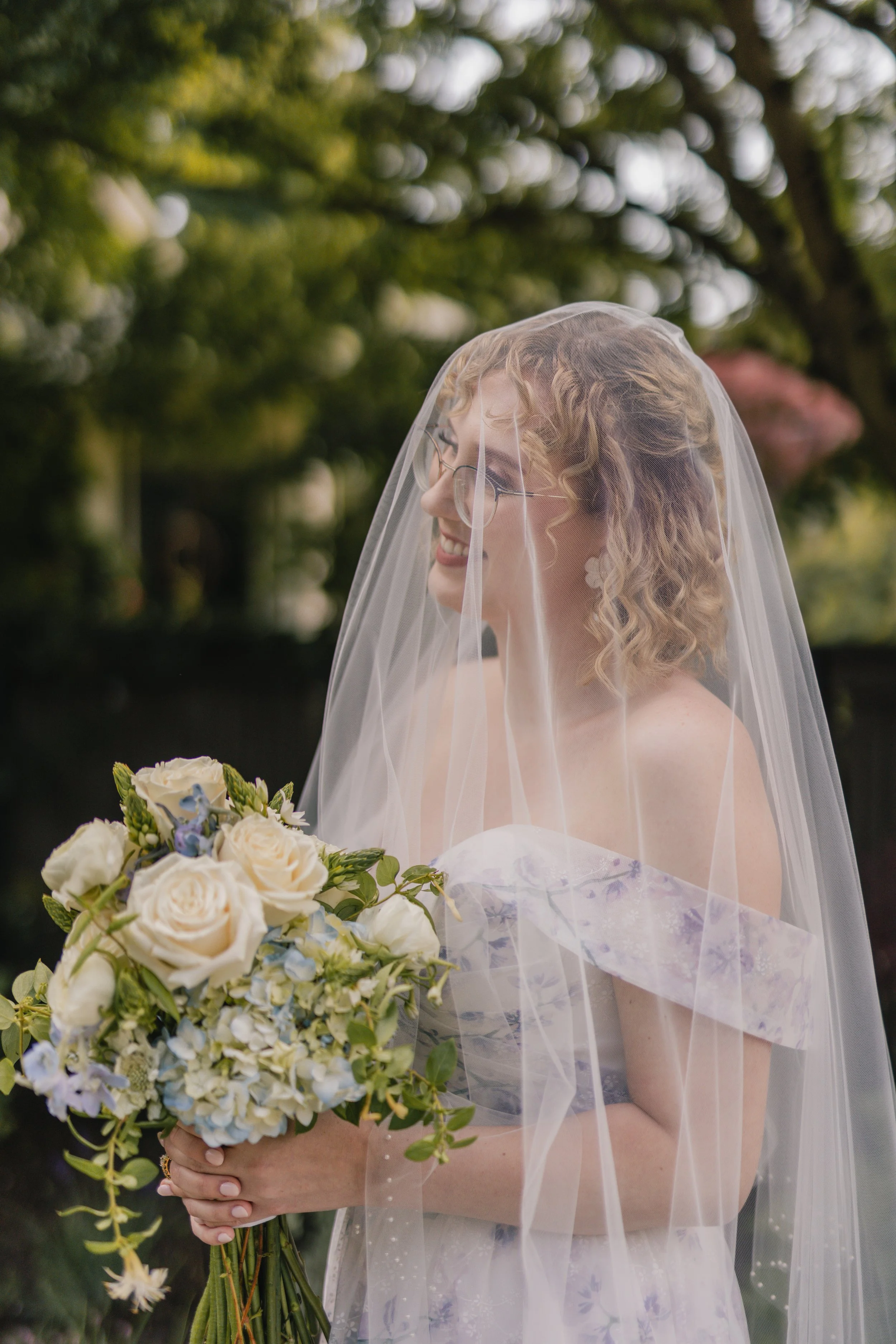 A woman with curly blonde hair, glasses, and a wedding veil holding a bouquet of white and light purple flowers outdoors.