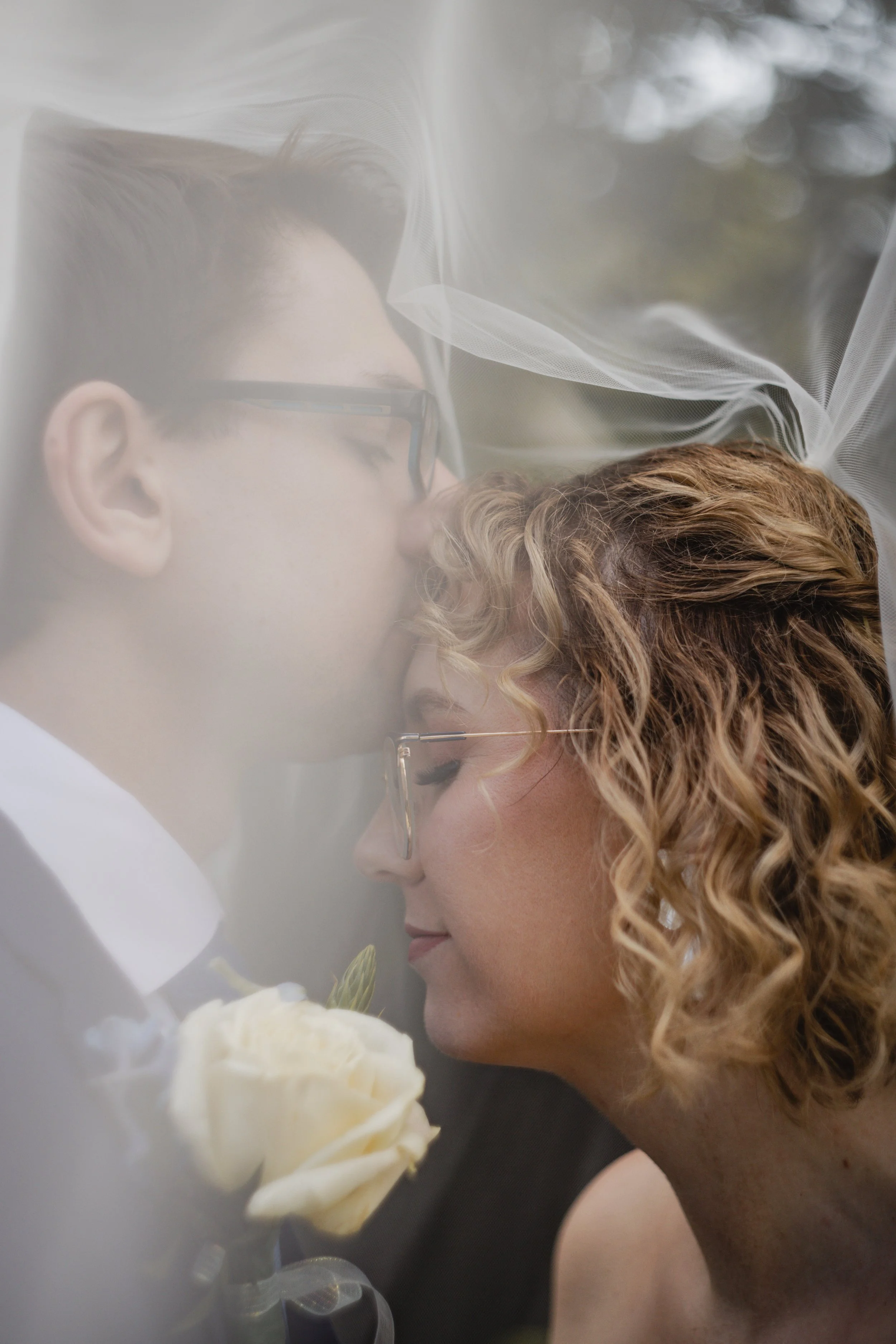 A bride with curly hair and glasses and a groom with short hair and glasses share a kiss through a sheer white veil at their wedding, with white flowers visible in the foreground.