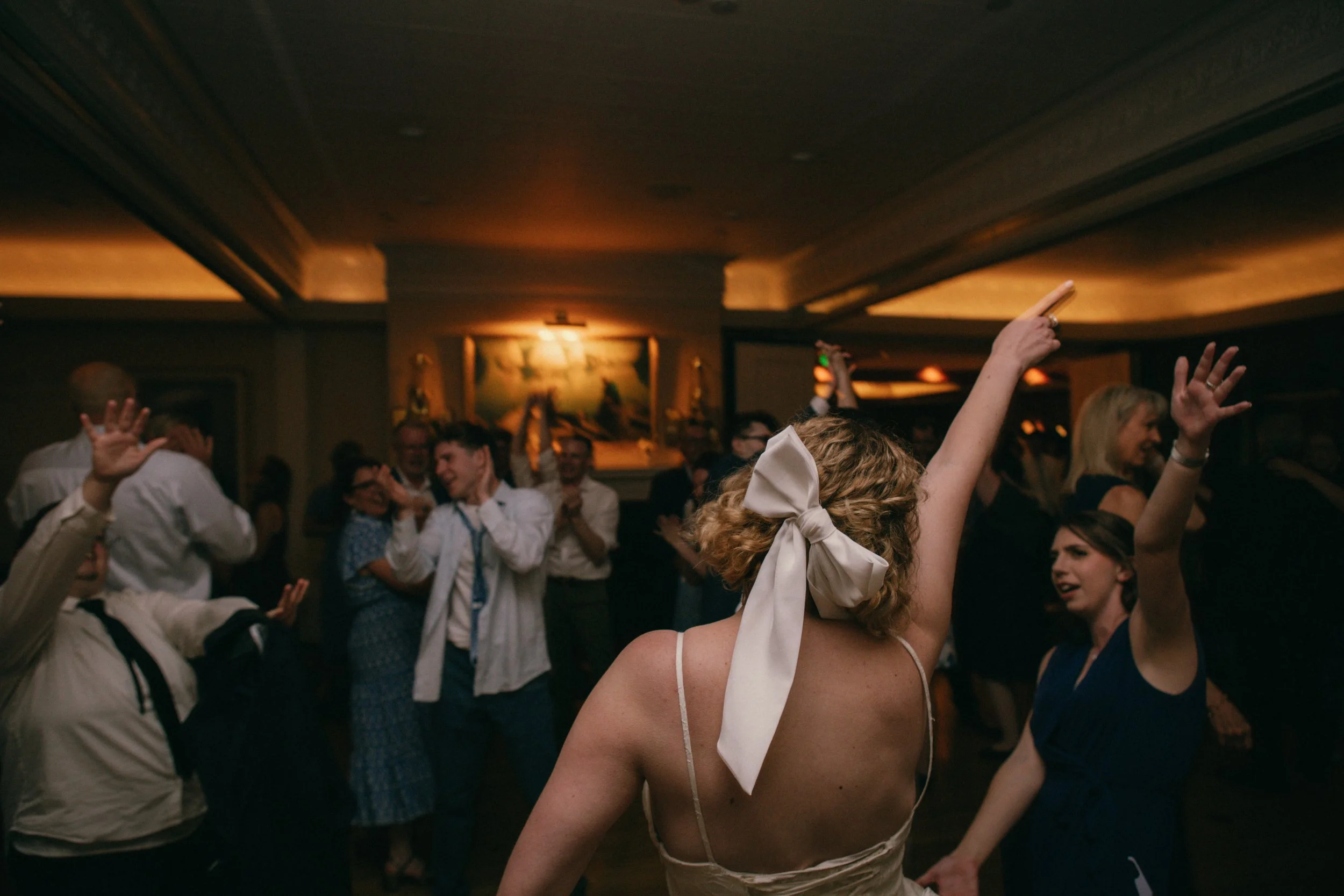 People dancing and enjoying an event in a dimly lit room with wooden ceiling. A woman with curly blonde hair, dressed in a beige slip dress with white ribbons, has her arms raised and is smiling, surrounded by others who are dancing and socializing.
