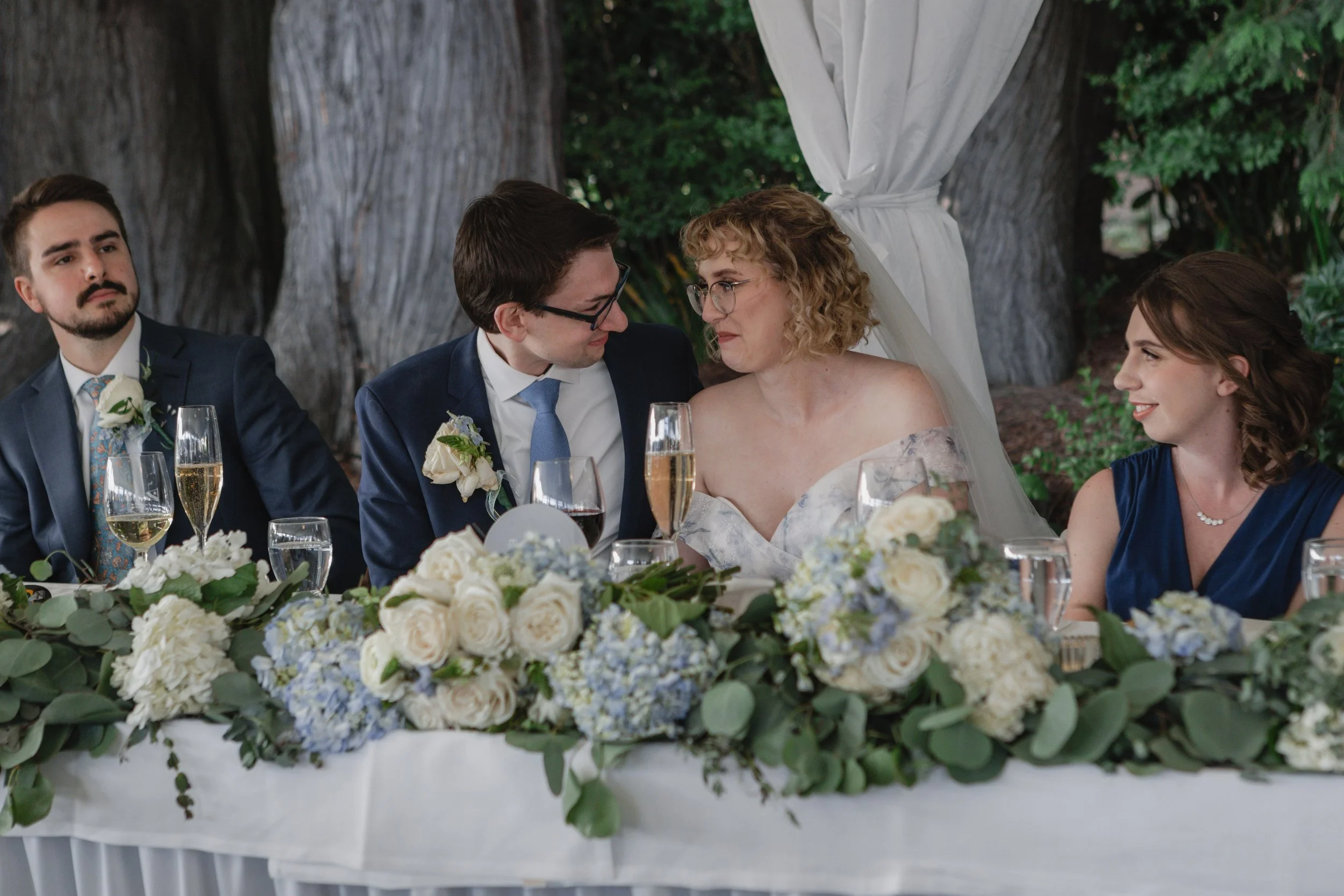 A wedding reception scene with a bride and groom sitting at a table decorated with white and blue flowers, surrounded by friends, outdoors under a canopy with trees in the background.