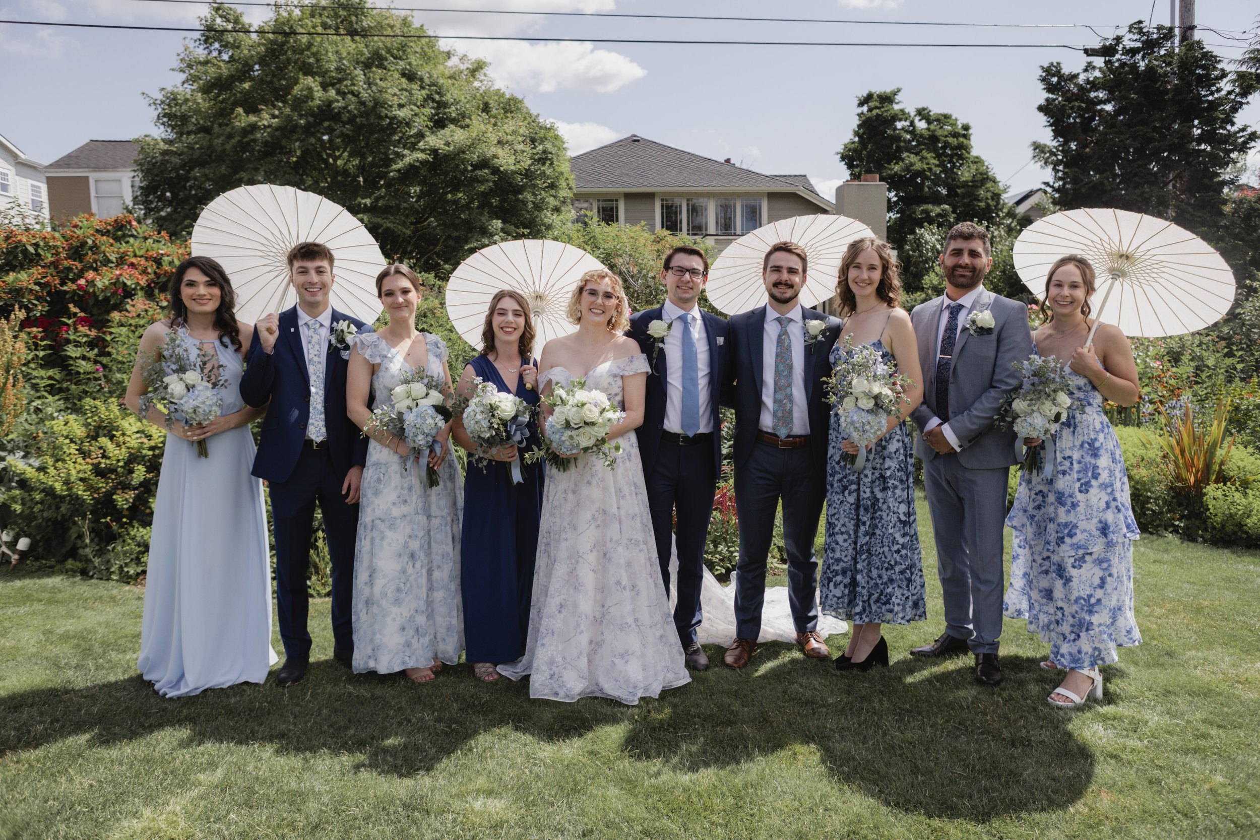 Group of people dressed in formal attire, holding umbrellas and bouquets, posing outdoors in a garden with a house and trees in the background.