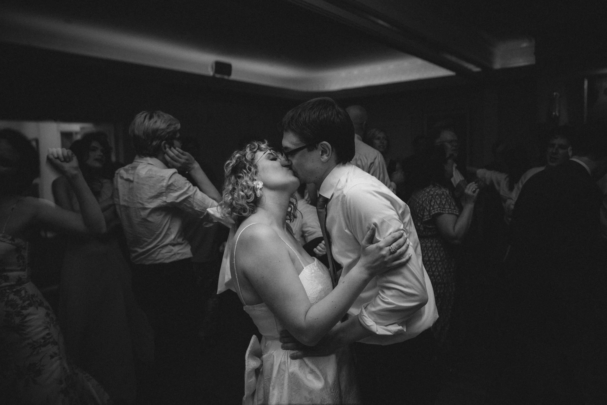 A black and white photo of a couple sharing a kiss at a celebration or party, with other guests in the background.