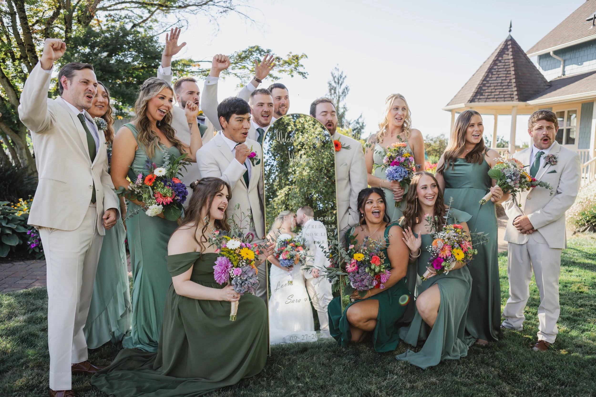 Group of people in wedding attire posing outdoors during a celebration, with colorful flower bouquets and a mirror with writing behind them.