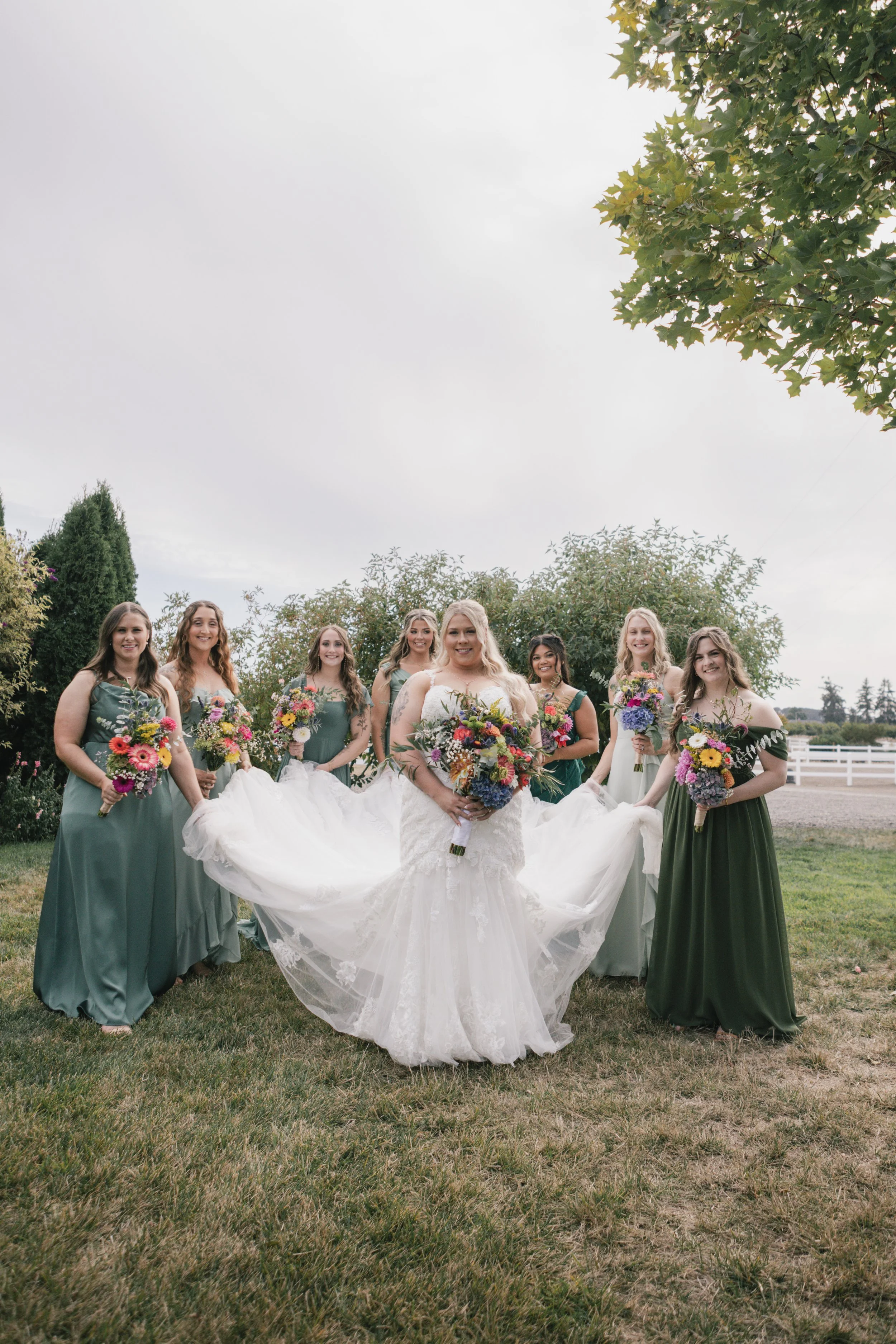 Group of eight women standing outdoors in a garden, dressed in formal attire with six women in matching green dresses and two in white dresses, holding colorful bouquets of flowers, under a large tree with green leaves on a cloudy day.