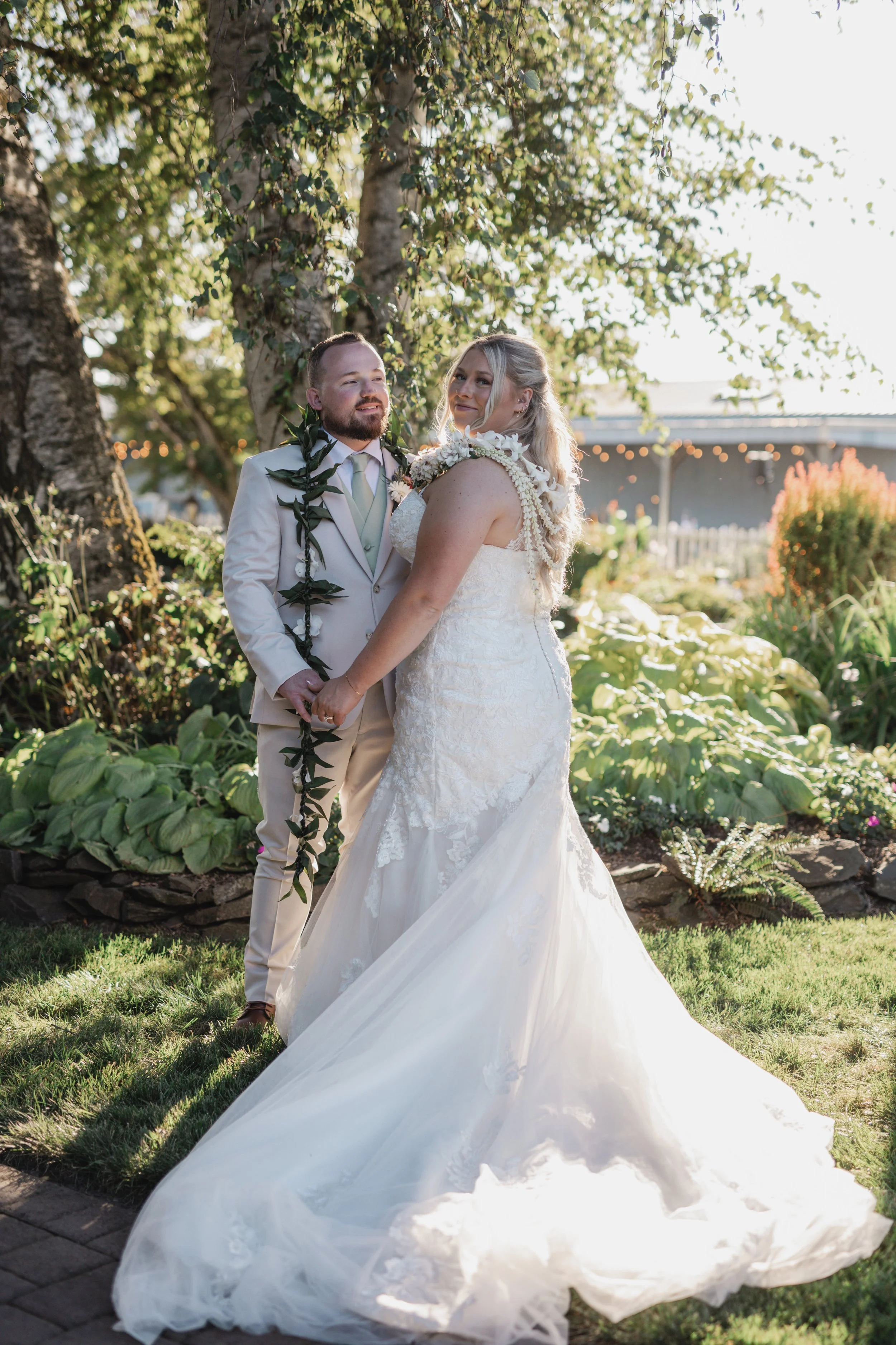 A bride and groom holding hands outdoors, standing on grass, with trees and a covered outdoor structure in the background. The groom wears a beige suit with a green tie, and the bride wears a white lace wedding dress with floral accents and a long tr