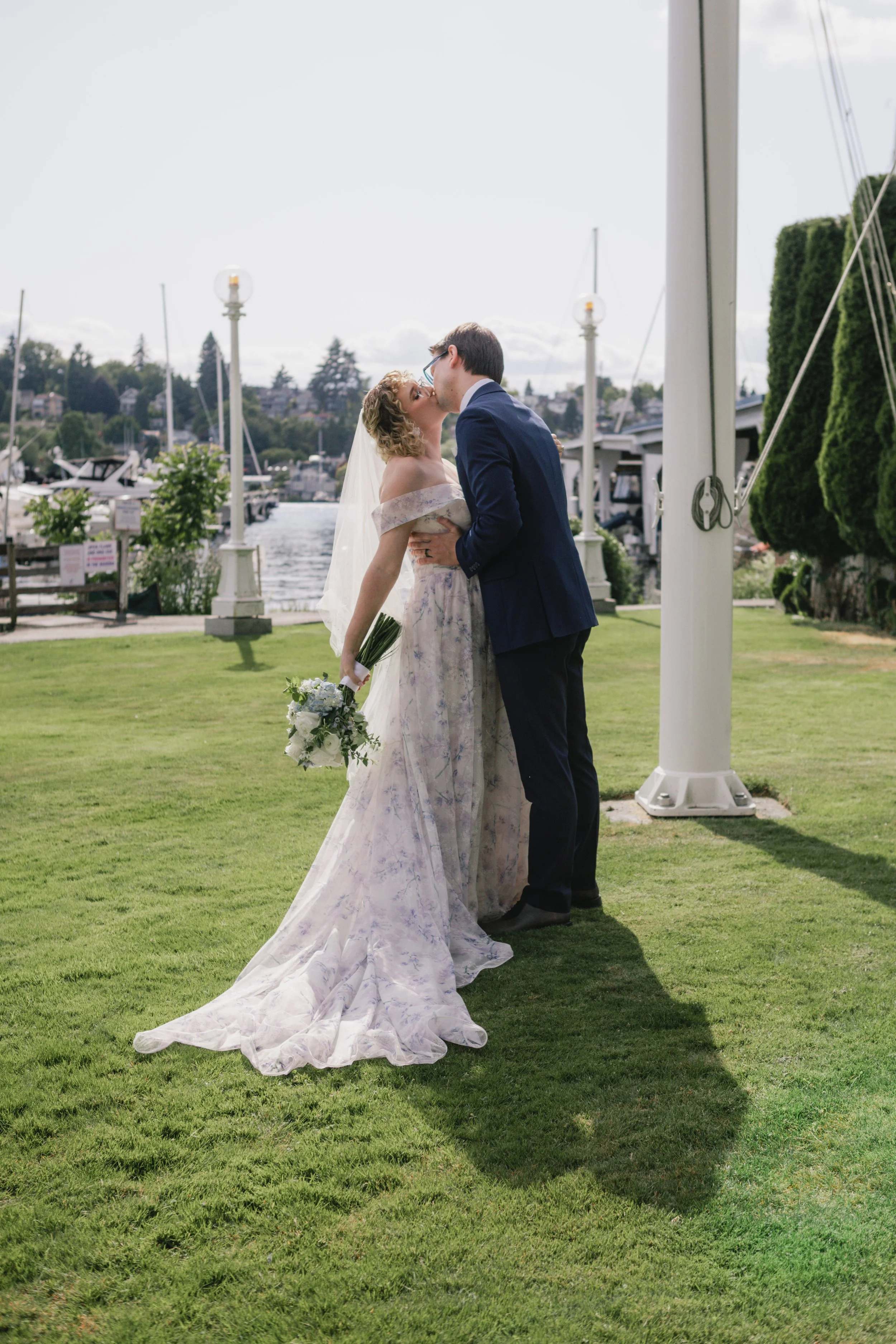 A couple kissing outdoors on a grassy lawn near a marina with boats, trees, and houses in the background. The woman is wearing a wedding dress and holding a bouquet, and the man is in a blue suit.