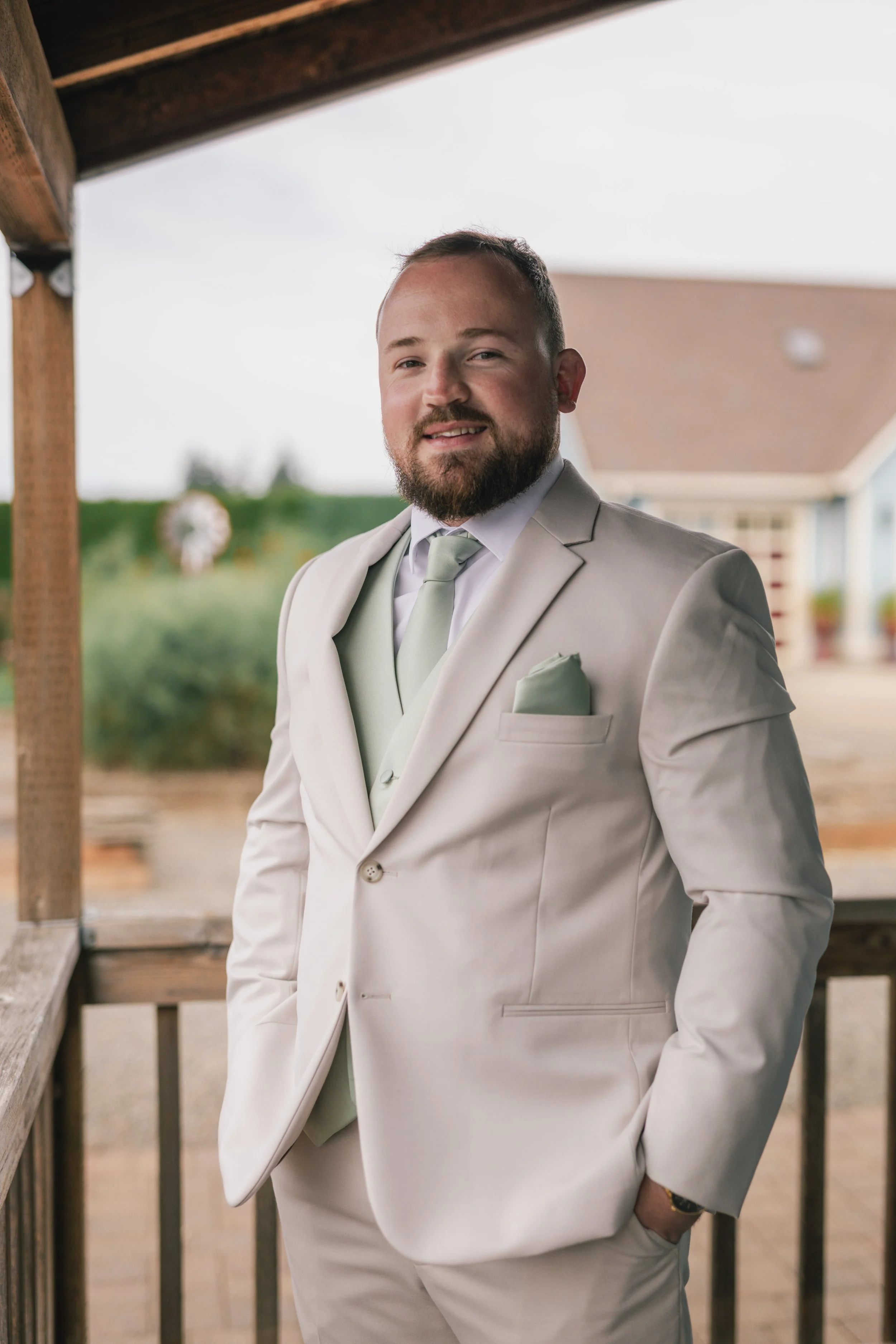 A man in a beige suit, light-colored tie, and pocket square standing outdoors beneath a wooden structure with a blurred background of greenery and a building.