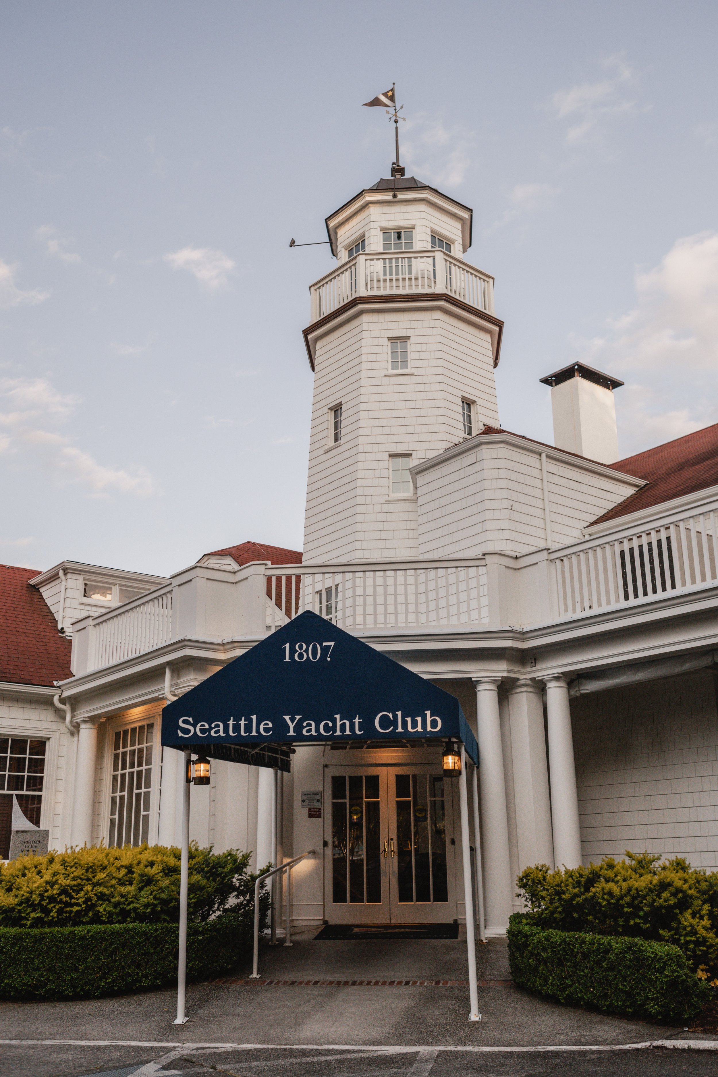 Front view of the Seattle Yacht Club building with a white exterior, a tower with a flag, and a blue canopy with the club's name. The entrance is flanked by neatly trimmed bushes and columns.