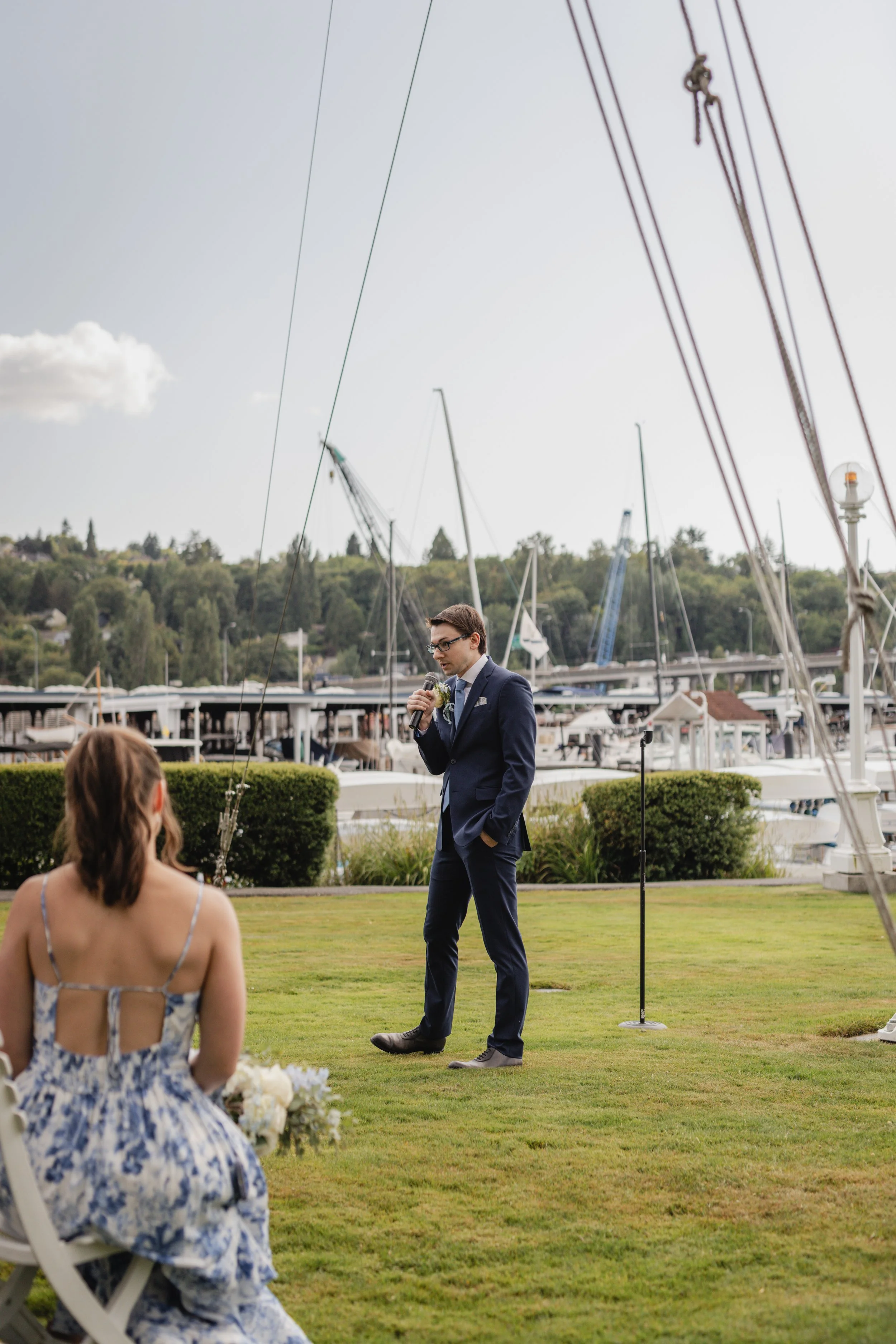 A man in a dark suit holding a microphone, standing on a grassy area with a marina and sailboats in the background. A woman in a floral dress is sitting in the foreground, facing the man.