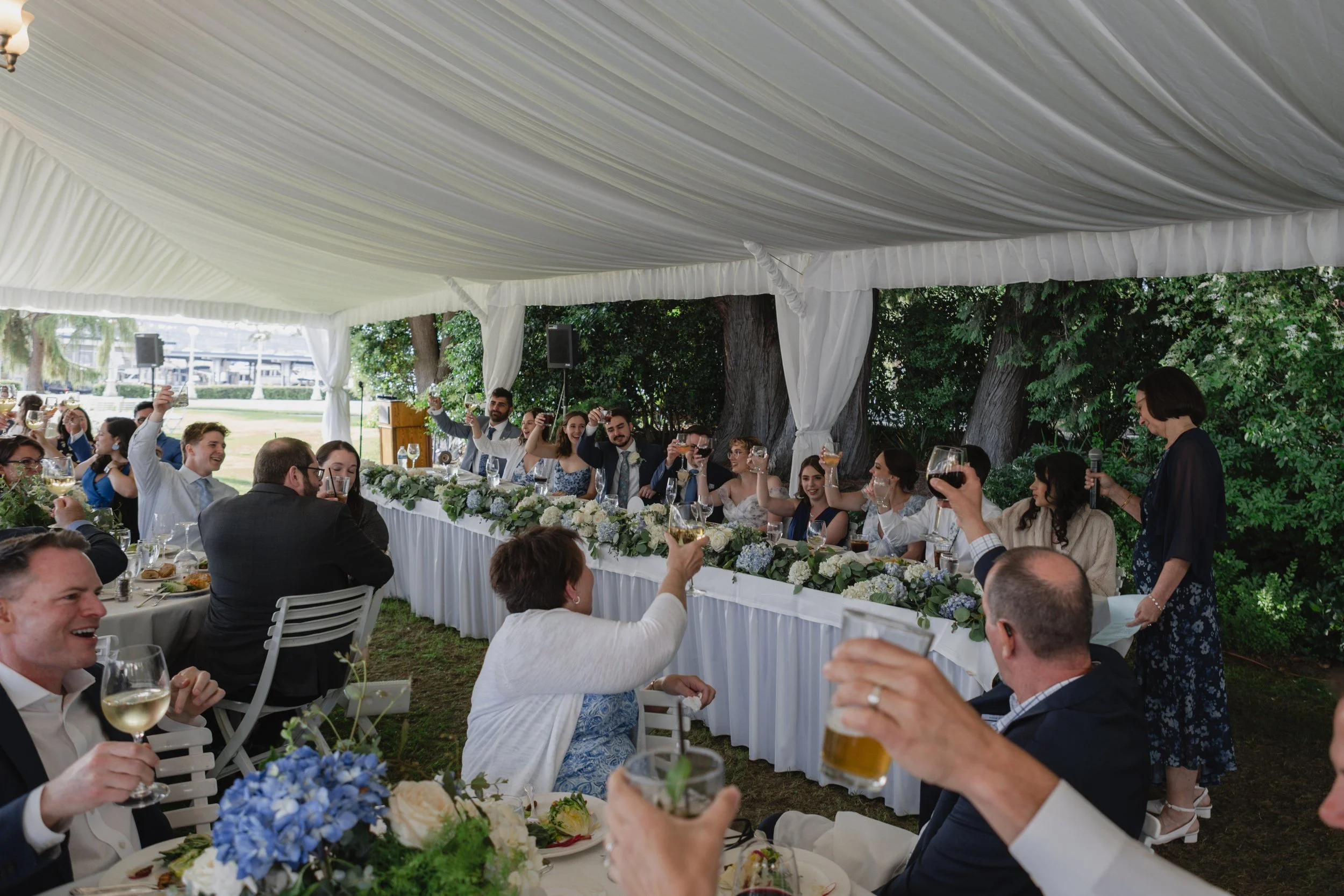 People attending a wedding reception outdoors, raising glasses in a toast under a decorated white canopy with trees in the background.