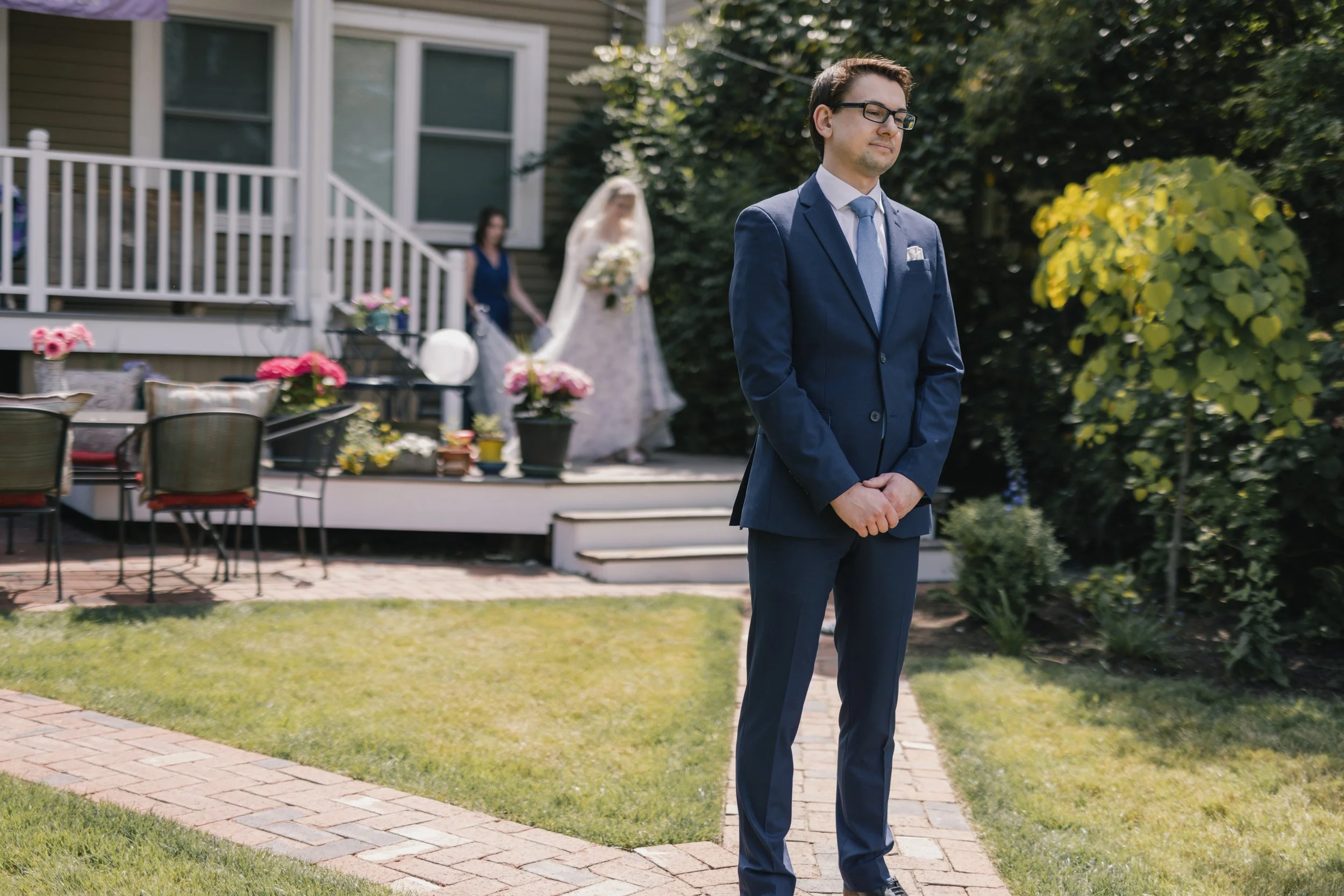 A man in a navy suit, light blue tie, and glasses stands with his hands clasped in front of him, outdoors in a garden, with a house and flower arrangements in the background. A bride in a white wedding dress and veil, holding flowers, is blurred in t