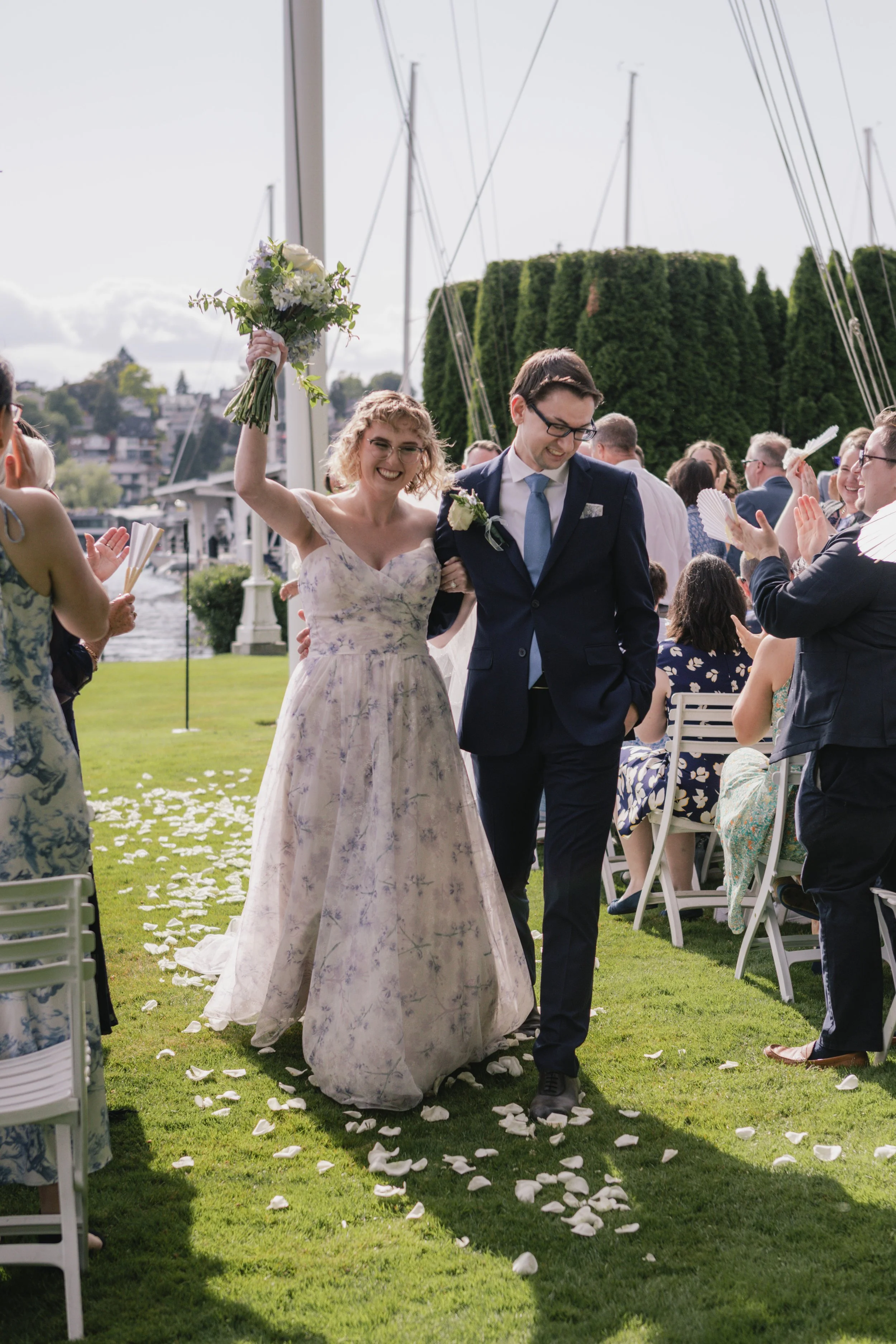 A bride and groom walking arm in arm down an aisle lined with white flower petals on grass, surrounded by seated guests clapping, with a marina and sailboats in the background.