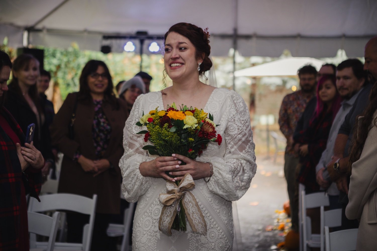 Bride in white dress holding bouquet, walking down aisle at wedding ceremony with seated guests.
