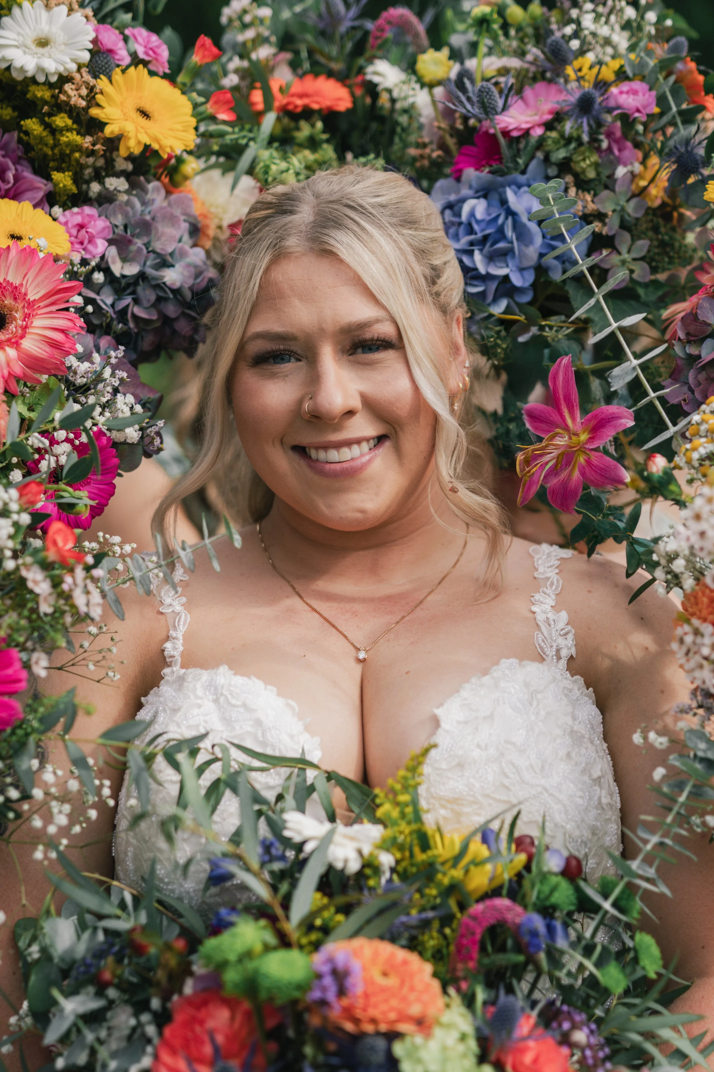 A smiling woman in a wedding dress surrounded by colorful flowers and greenery.