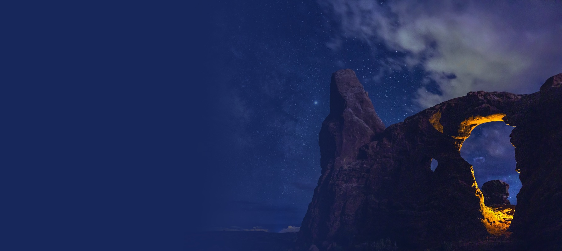 A towering sandstone arch lit warm gold at night, beneath a sky of stars and drifting cloud, fading into deep blue.